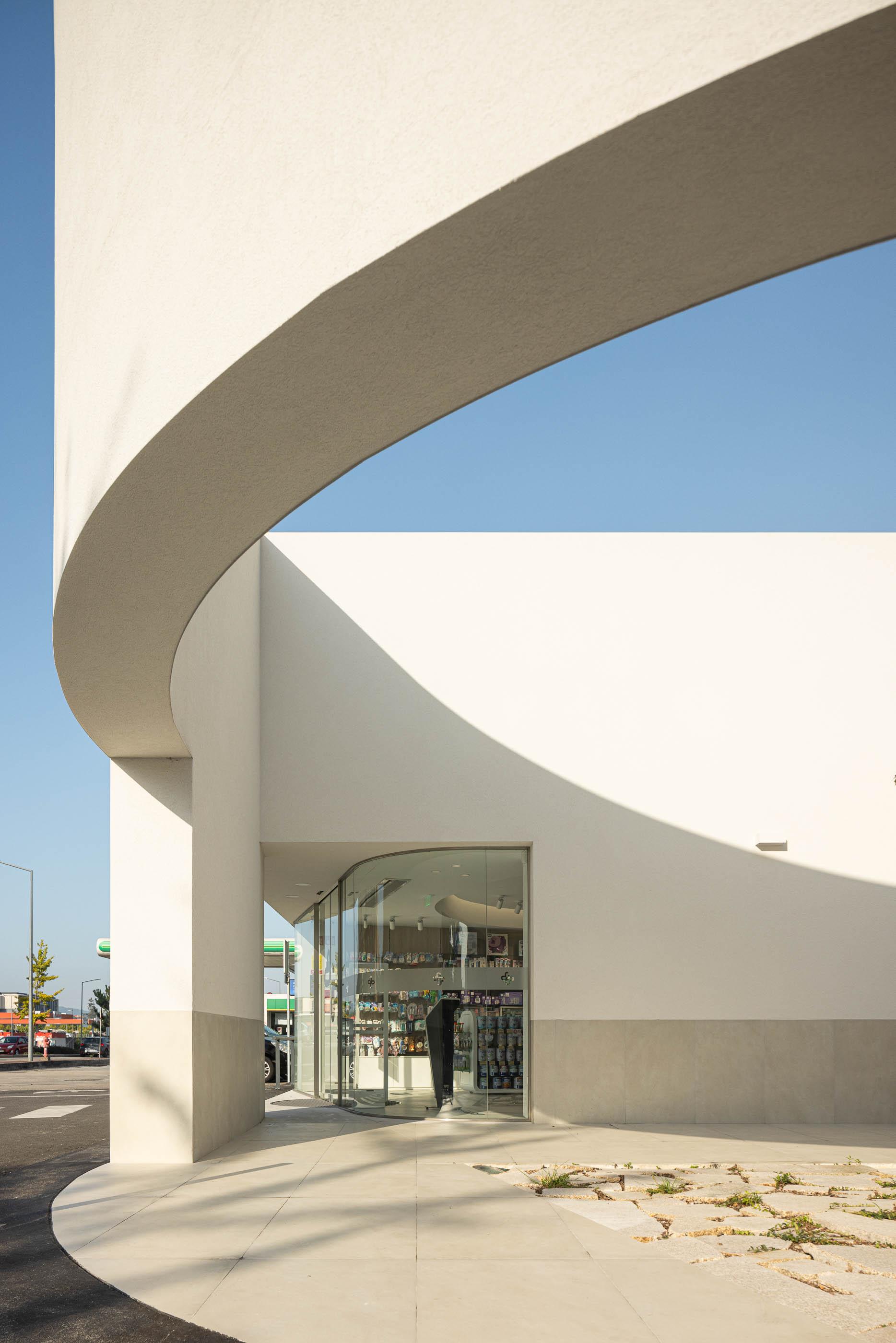 Exterior: Detalhe da fachada curva com entrada envidraçada e pavimento exterior em pedra. — Farmácia da Devesa arquitetura Tsou Arquitectos em Vila Nova de Famalicão