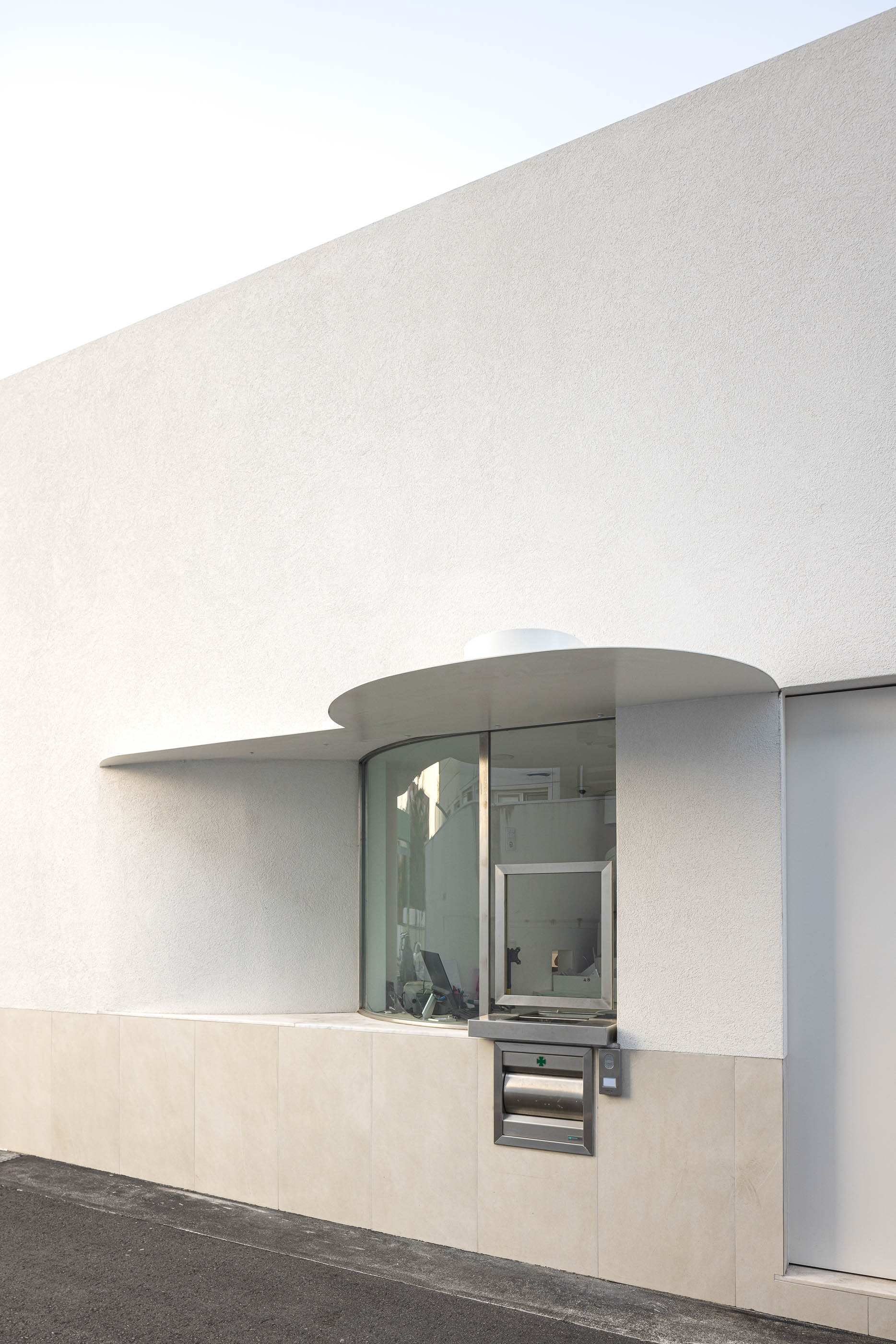 Exterior: Fachada curva branca com janela de atendimento "Drive Farma" e estacionamento. — Farmácia da Devesa arquitetura Tsou Arquitectos em Vila Nova de Famalicão