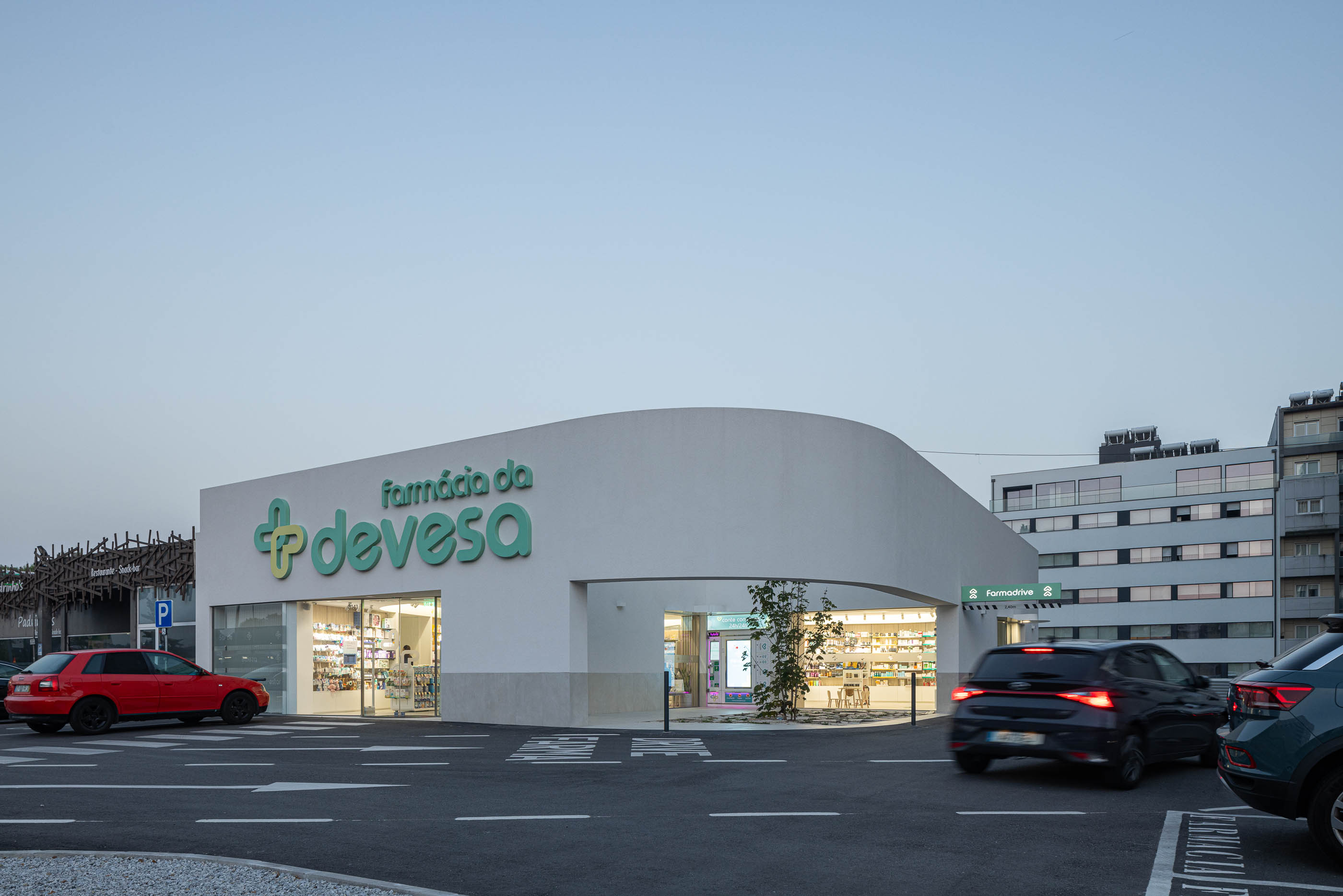 Exterior: Vista noturna da farmácia com janela de atendimento "Drive Farma" e jardim interior iluminado. — Farmácia da Devesa arquitetura Tsou Arquitectos em Vila Nova de Famalicão
