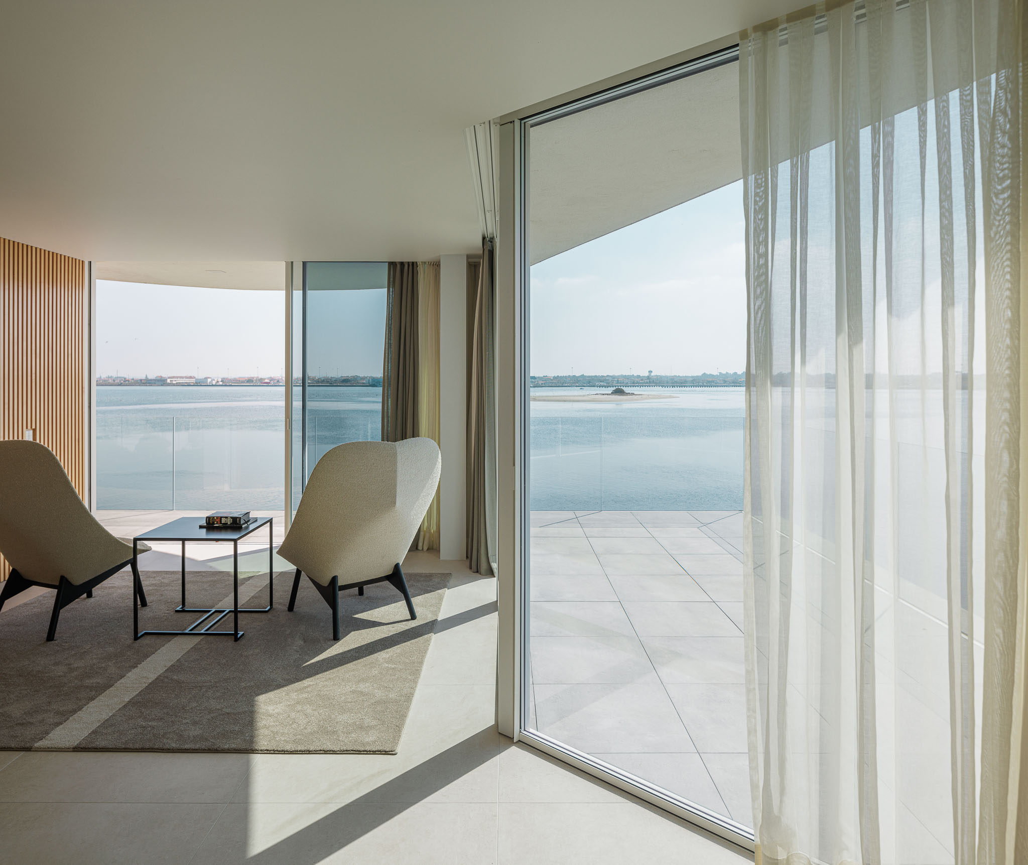 Sala de estar: Cadeirões modernos, mesa de apoio preta, alcatifa cinzenta e vista panorâmica para a ria através de grandes janelas. — Moradia JD2 arquitetura Rui Rosmaninho Arquiteto em Ílhavo
