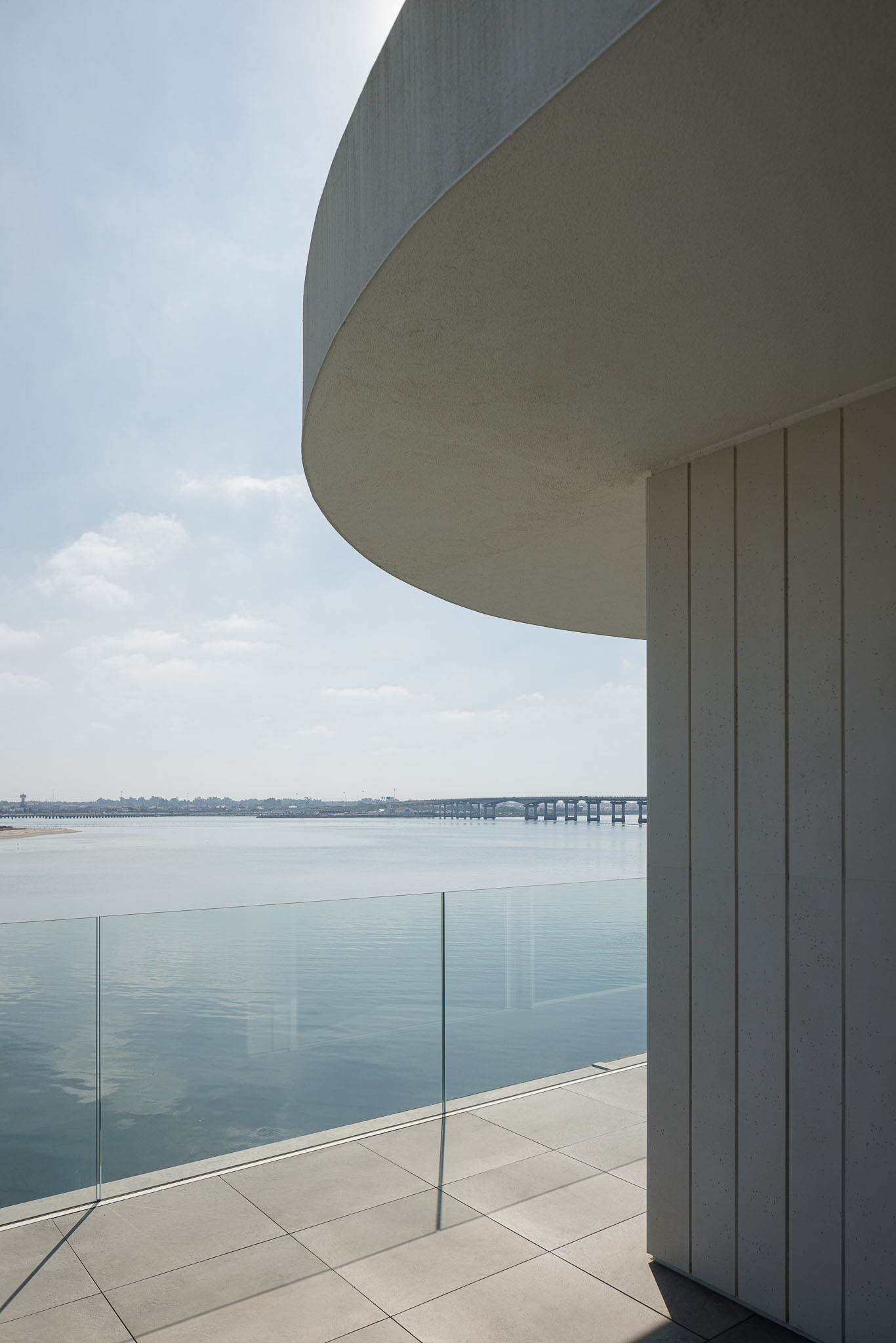 Varanda: Pavimento cerâmico cinzento, guarda em vidro e vista para a ria, com elementos arquitetónicos curvos e revestimento vertical. — Moradia JD2 arquitetura Rui Rosmaninho Arquiteto em Ílhavo
