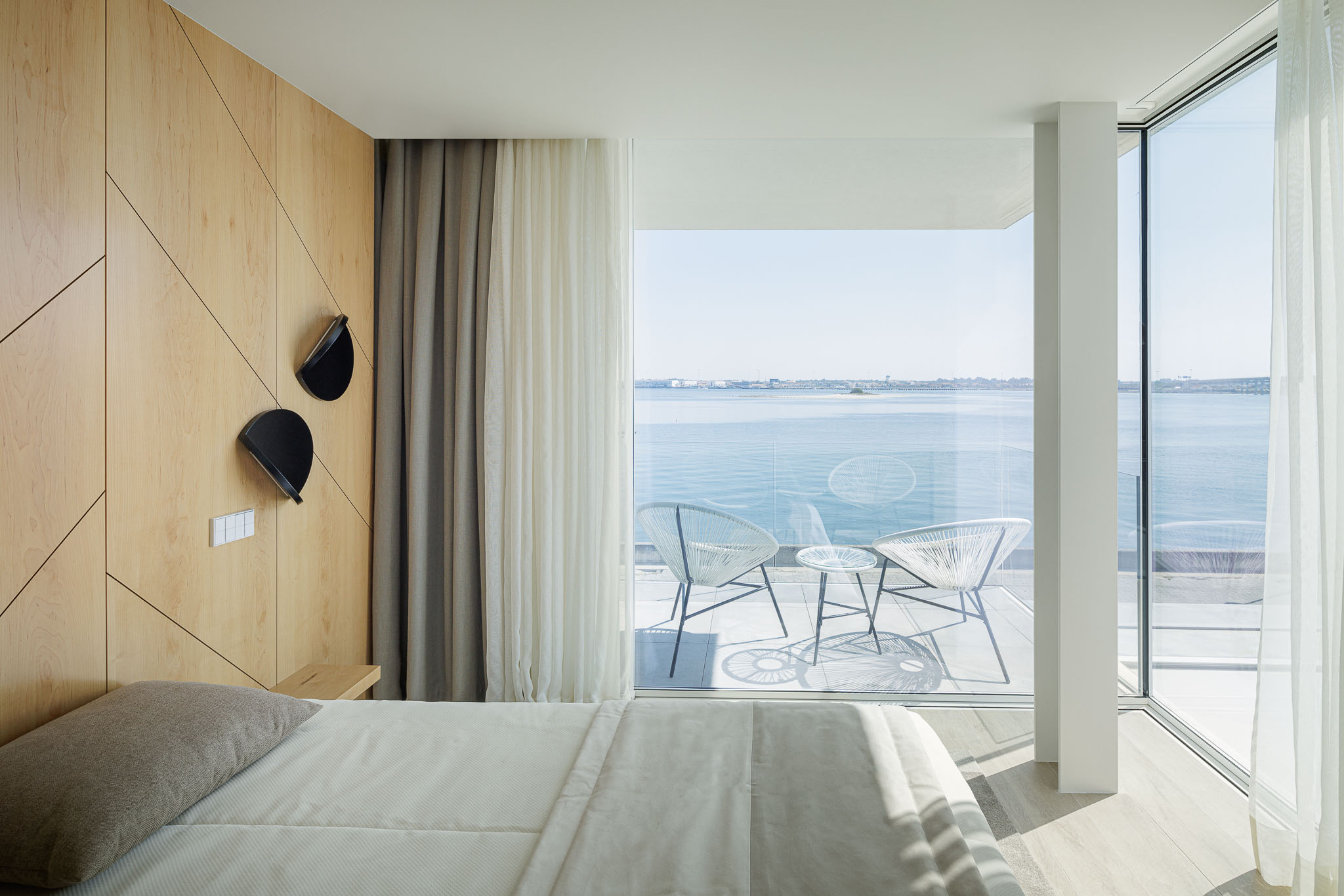 Quarto: Cama com cabeceira em madeira, candeeiros de parede pretos e acesso à varanda com vista para a ria. — Moradia JD2 arquitetura Rui Rosmaninho Arquiteto em Ílhavo