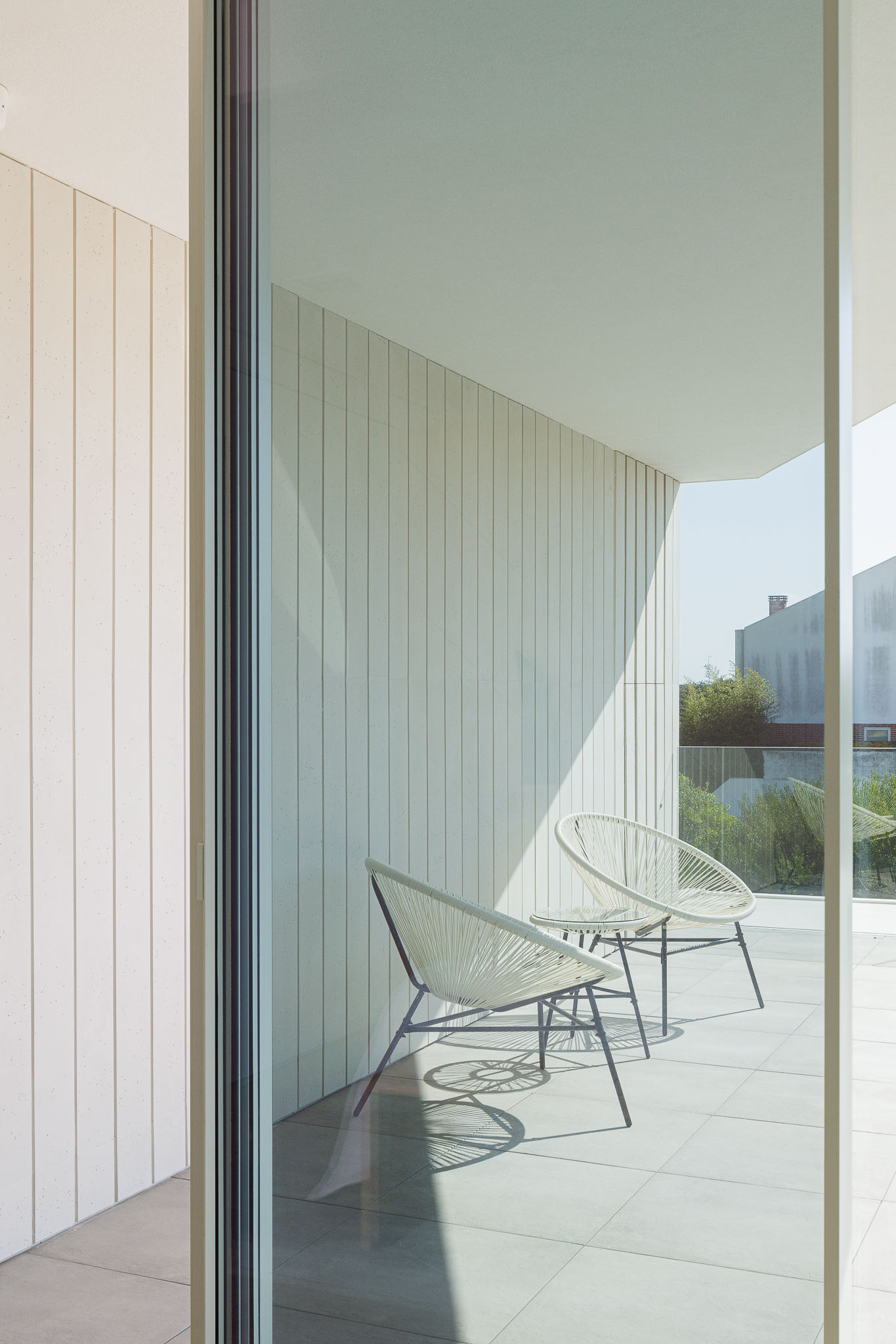 Varanda: Cadeiras de vime brancas, pavimento cerâmico cinzento e revestimento vertical branco. — Moradia JD2 arquitetura Rui Rosmaninho Arquiteto em Ílhavo