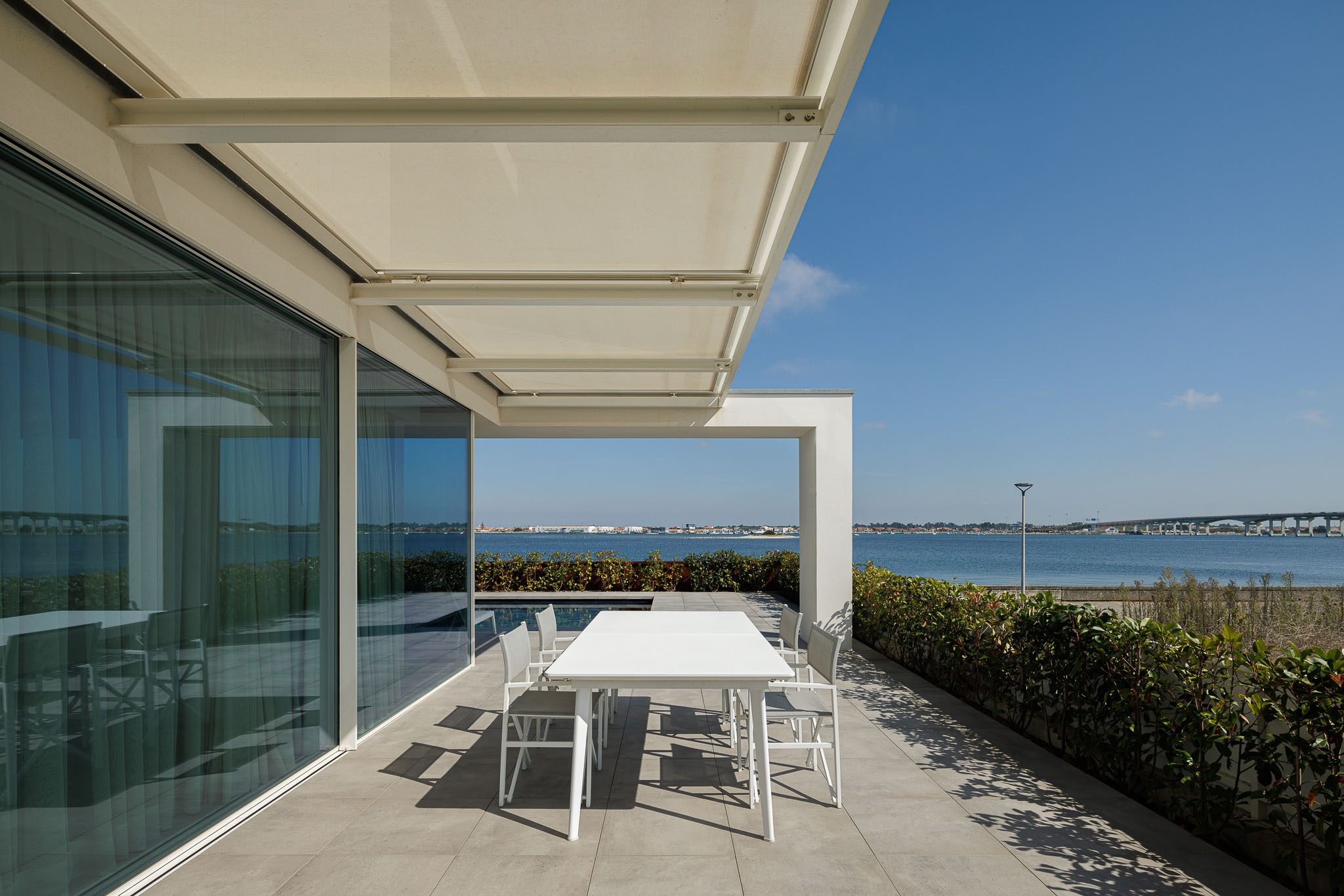 Alpendre: Mesa e cadeiras brancas, pavimento cerâmico cinzento e vista para a piscina e ria. — Moradia JD2 arquitetura Rui Rosmaninho Arquiteto em Ílhavo