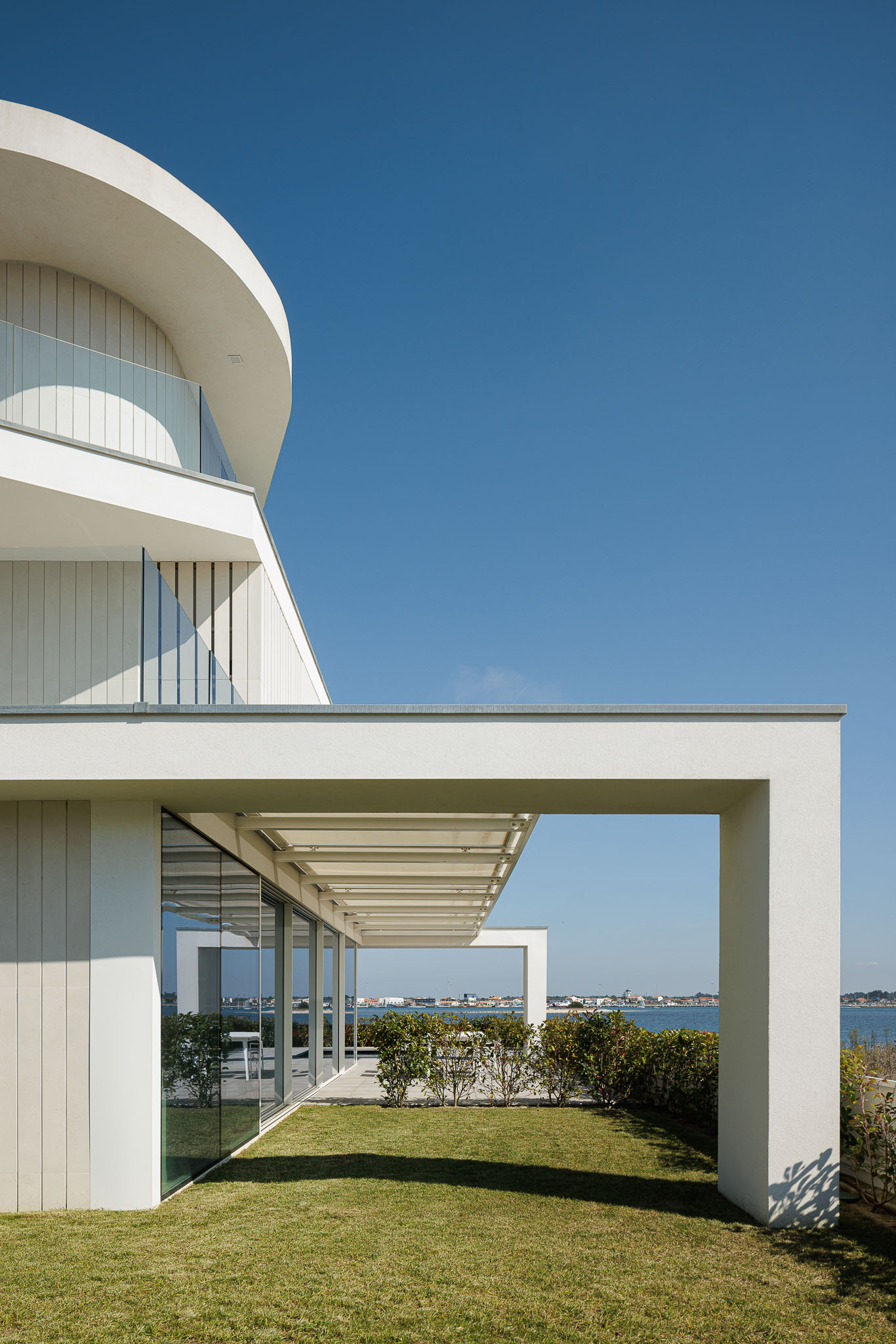 Exterior: Fachada branca com elementos curvos, varandas com guarda em vidro e jardim relvado. — Moradia JD2 arquitetura Rui Rosmaninho Arquiteto em Ílhavo