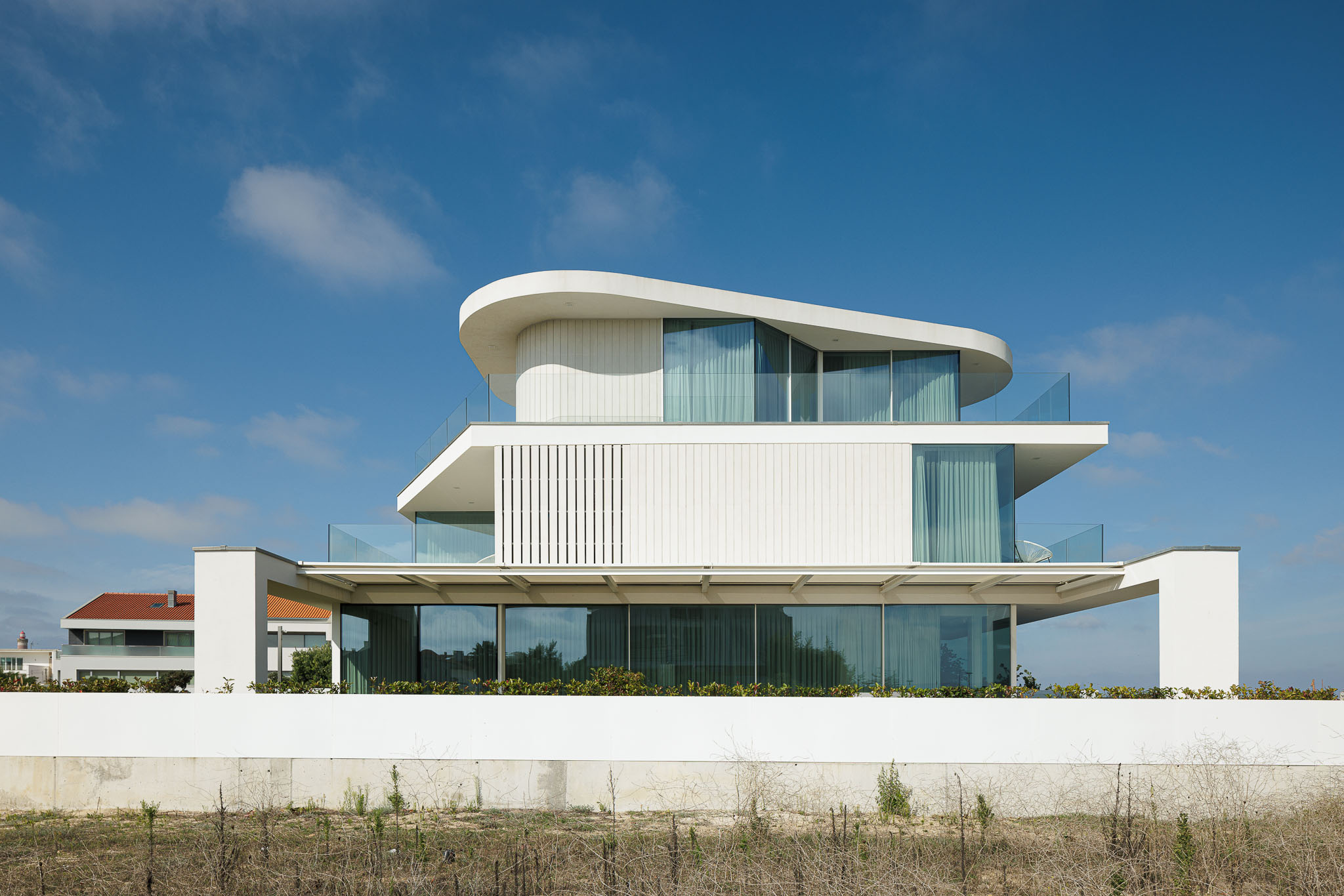 Exterior: Vista geral da moradia com fachada branca, varandas com guarda em vidro e cobertura curva. — Moradia JD2 arquitetura Rui Rosmaninho Arquiteto em Ílhavo