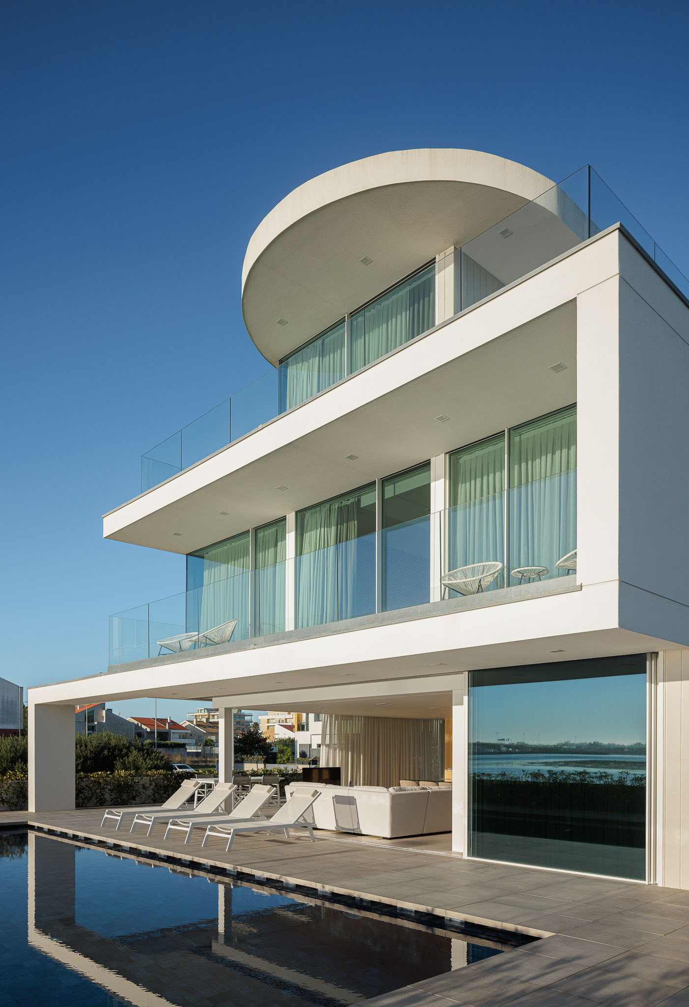 Exterior: Piscina com espreguiçadeiras brancas, pavimento cerâmico cinzento e vista para a ria. — Moradia JD2 arquitetura Rui Rosmaninho Arquiteto em Ílhavo
