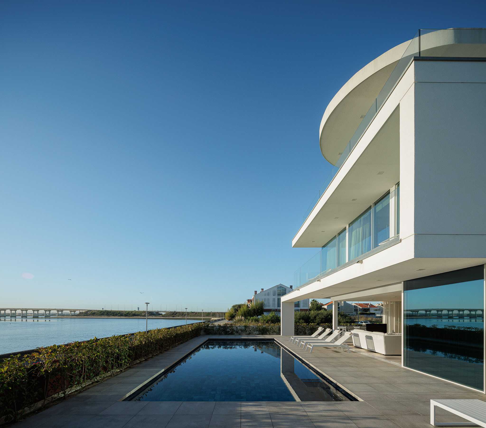 Sala de estar: Sofá branco, mesa de jantar com tampo de vidro e cadeiras cinzentas, e vista para a piscina. — Moradia JD2 arquitetura Rui Rosmaninho Arquiteto em Ílhavo