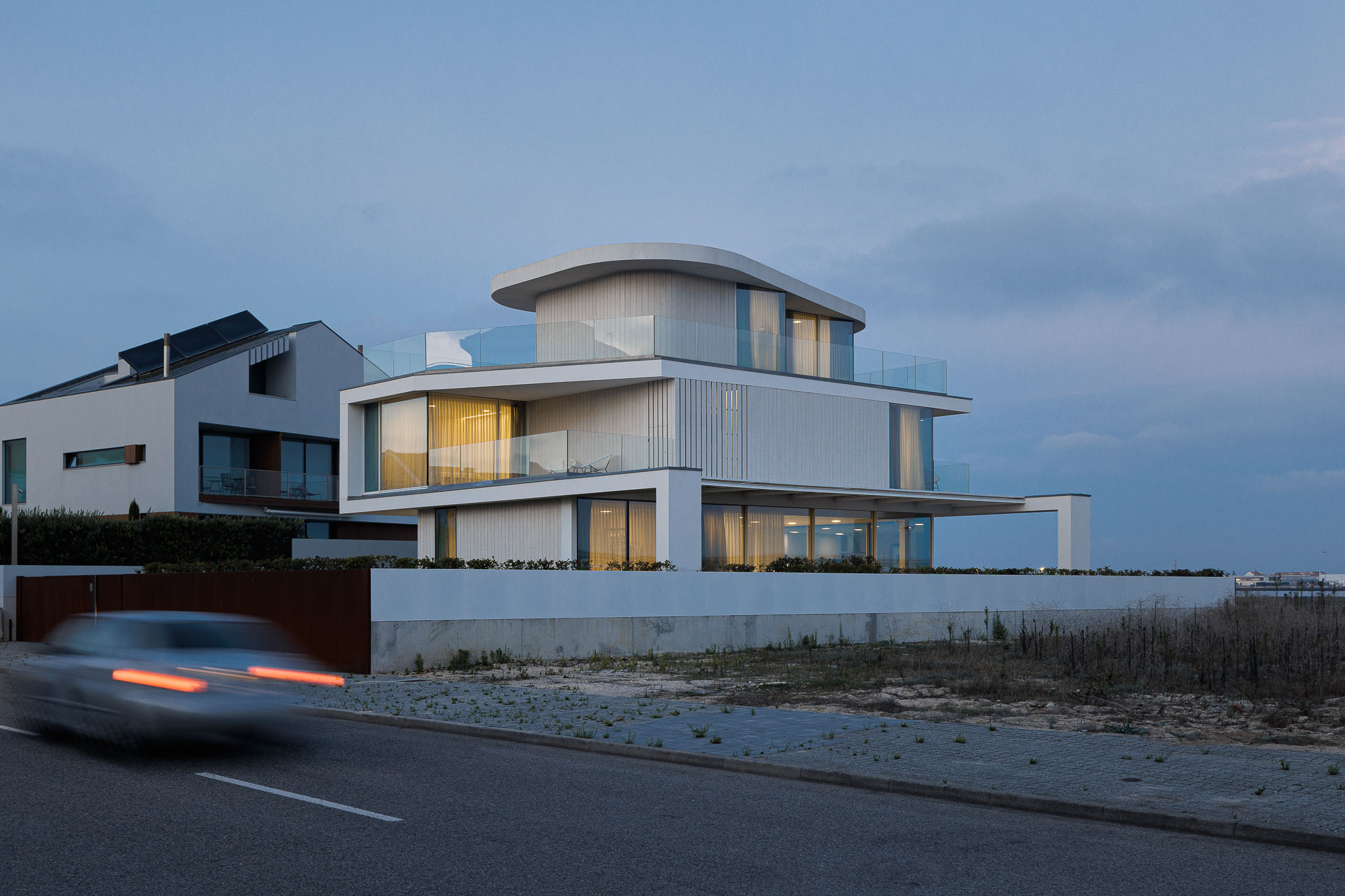 Exterior: Moradia moderna iluminada ao anoitecer, com fachada branca e varandas com guarda em vidro. — Moradia JD2 arquitetura Rui Rosmaninho Arquiteto em Ílhavo