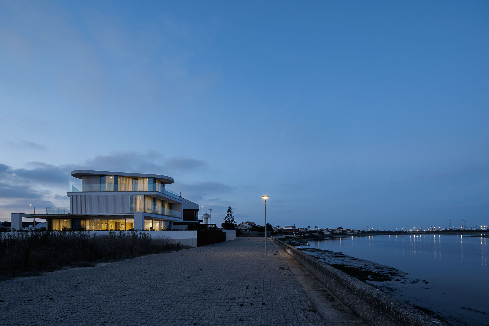 Exterior: Moradia moderna iluminada ao anoitecer, com fachada branca e varandas com guarda em vidro. — Moradia JD2 arquitetura Rui Rosmaninho Arquiteto em Ílhavo