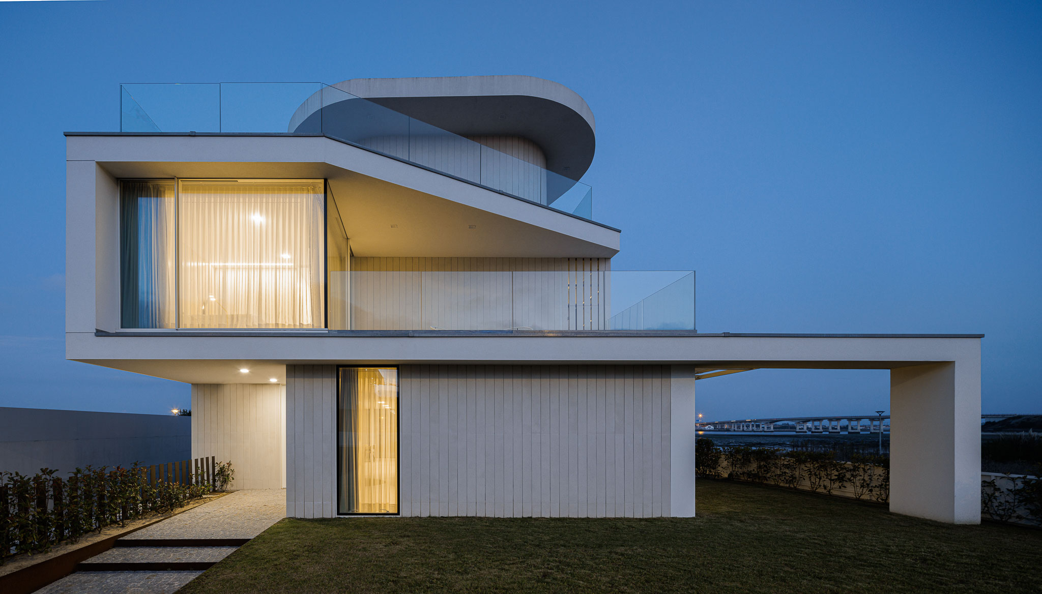 Exterior: Detalhe da fachada com elementos curvos, varandas com guarda em vidro e iluminação interior. — Moradia JD2 arquitetura Rui Rosmaninho Arquiteto em Ílhavo