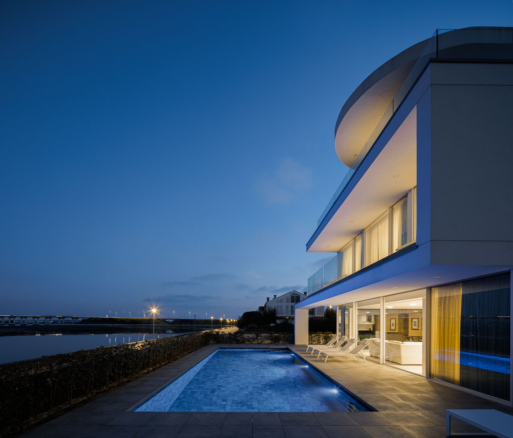 Exterior: Detalhe da varanda com pavimento cerâmico cinzento, guarda em vidro e vista para a ria ao anoitecer. — Moradia JD2 arquitetura Rui Rosmaninho Arquiteto em Ílhavo