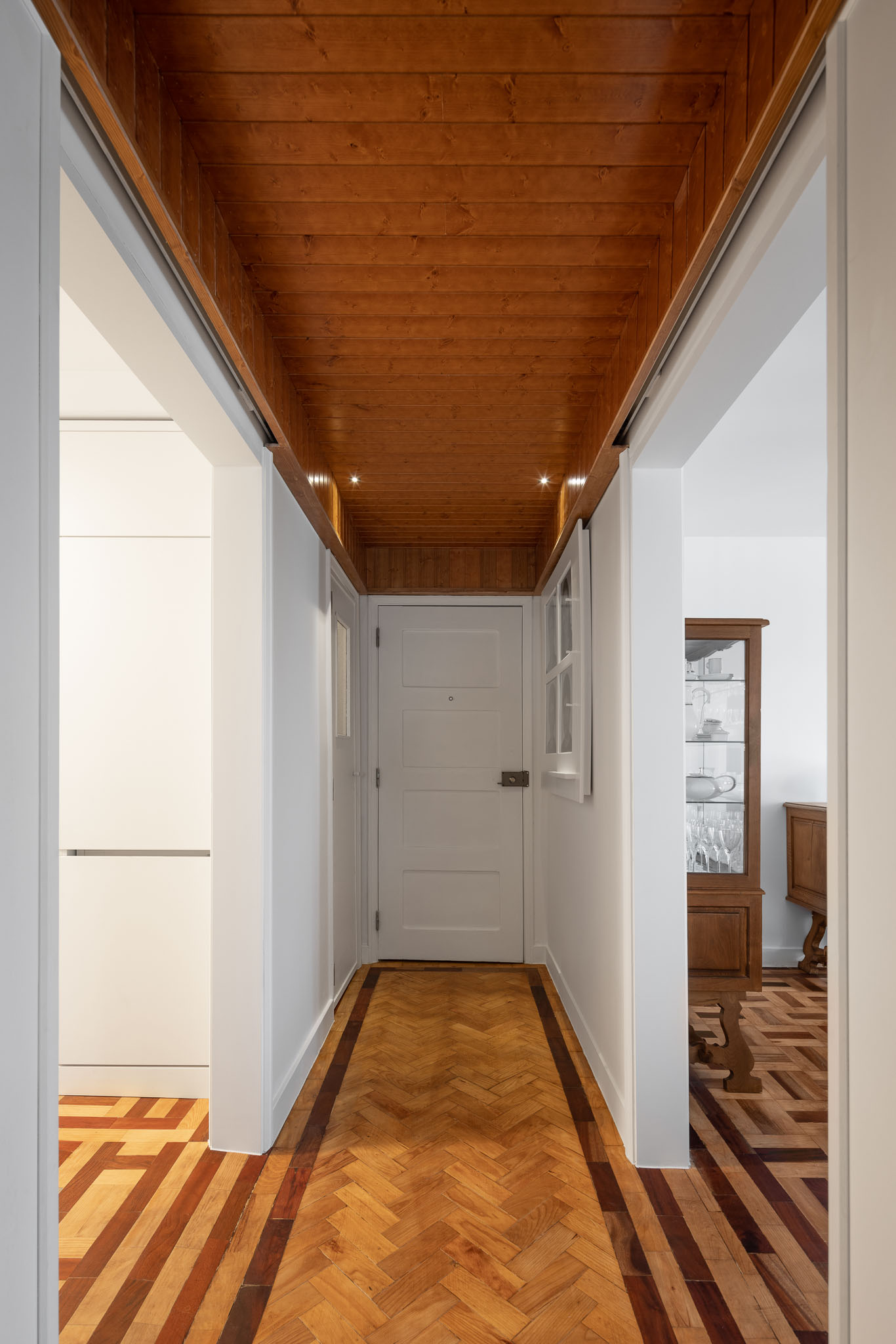 Corredor: Pavimento em madeira com padrão geométrico, teto em madeira e paredes brancas. Porta de entrada ao fundo. — Apartamento em Gouveia arquitetura M2Senos Arquitectos em Gouveia