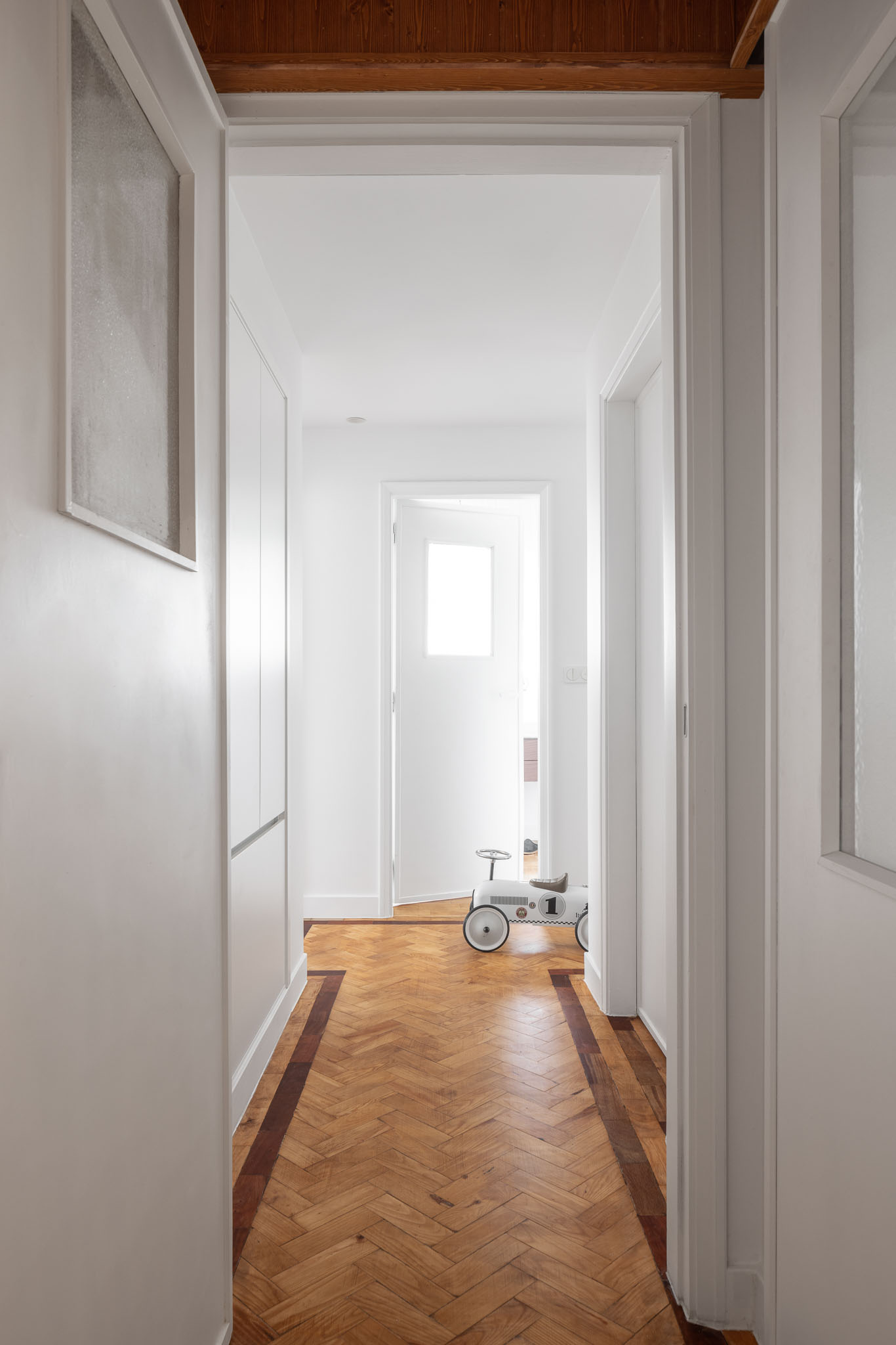 Corredor: Vista do corredor com pavimento em madeira, paredes brancas e portas brancas. Um triciclo infantil visível ao fundo. — Apartamento em Gouveia arquitetura M2Senos Arquitectos em Gouveia
