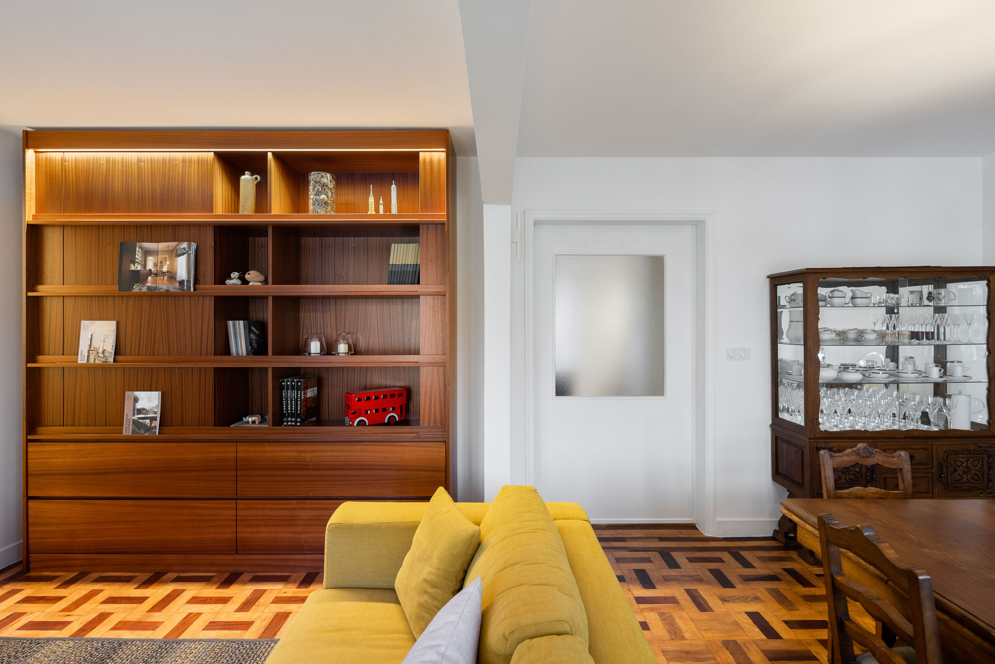Sala de estar: Estante em madeira com iluminação embutida, sofá amarelo e pavimento em madeira com padrão geométrico. — Apartamento em Gouveia arquitetura M2Senos Arquitectos em Gouveia