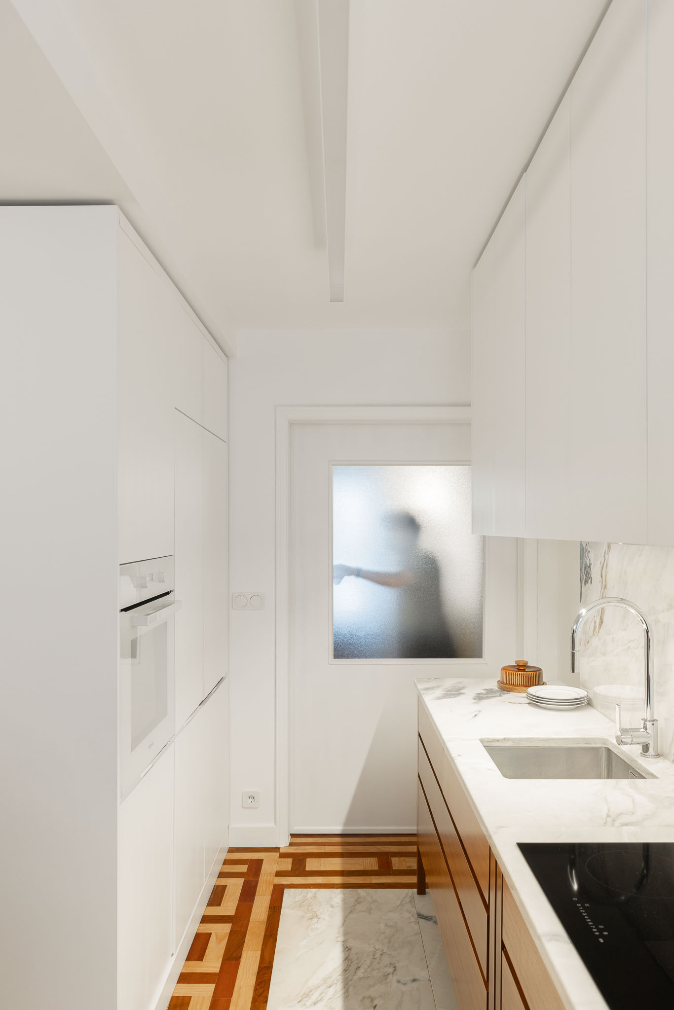 Cozinha: Vista geral da cozinha com armários brancos, bancada em mármore e pavimento em madeira. Porta com vidro fosco. — Apartamento em Gouveia arquitetura M2Senos Arquitectos em Gouveia
