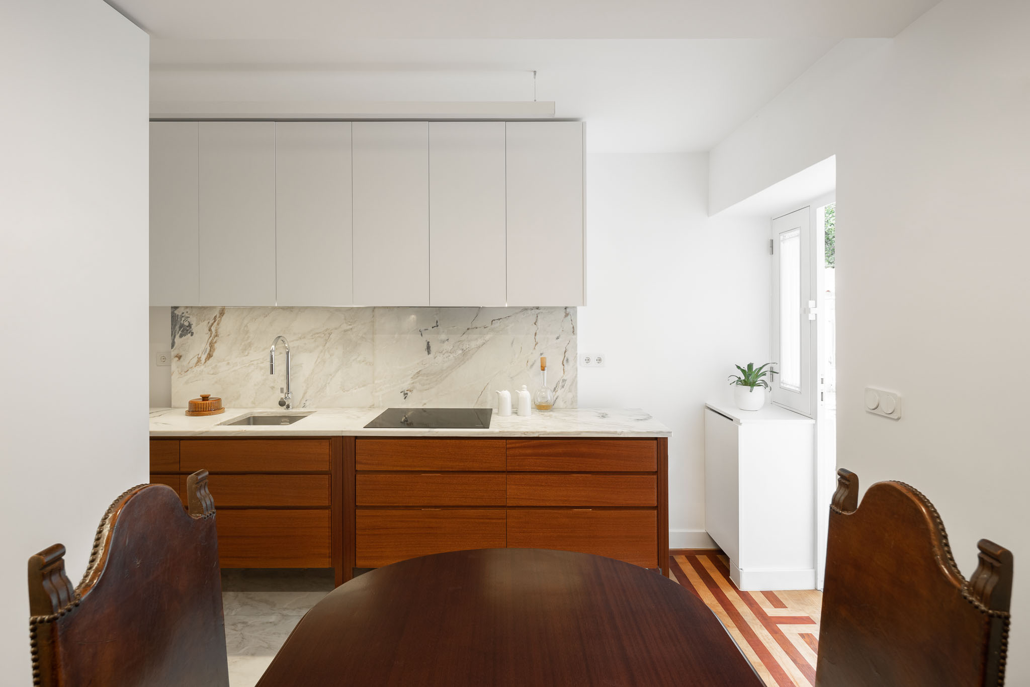 Cozinha: Perspectiva da cozinha com armários brancos, bancada em mármore e mesa de jantar ao fundo. Pavimento em madeira. — Apartamento em Gouveia arquitetura M2Senos Arquitectos em Gouveia