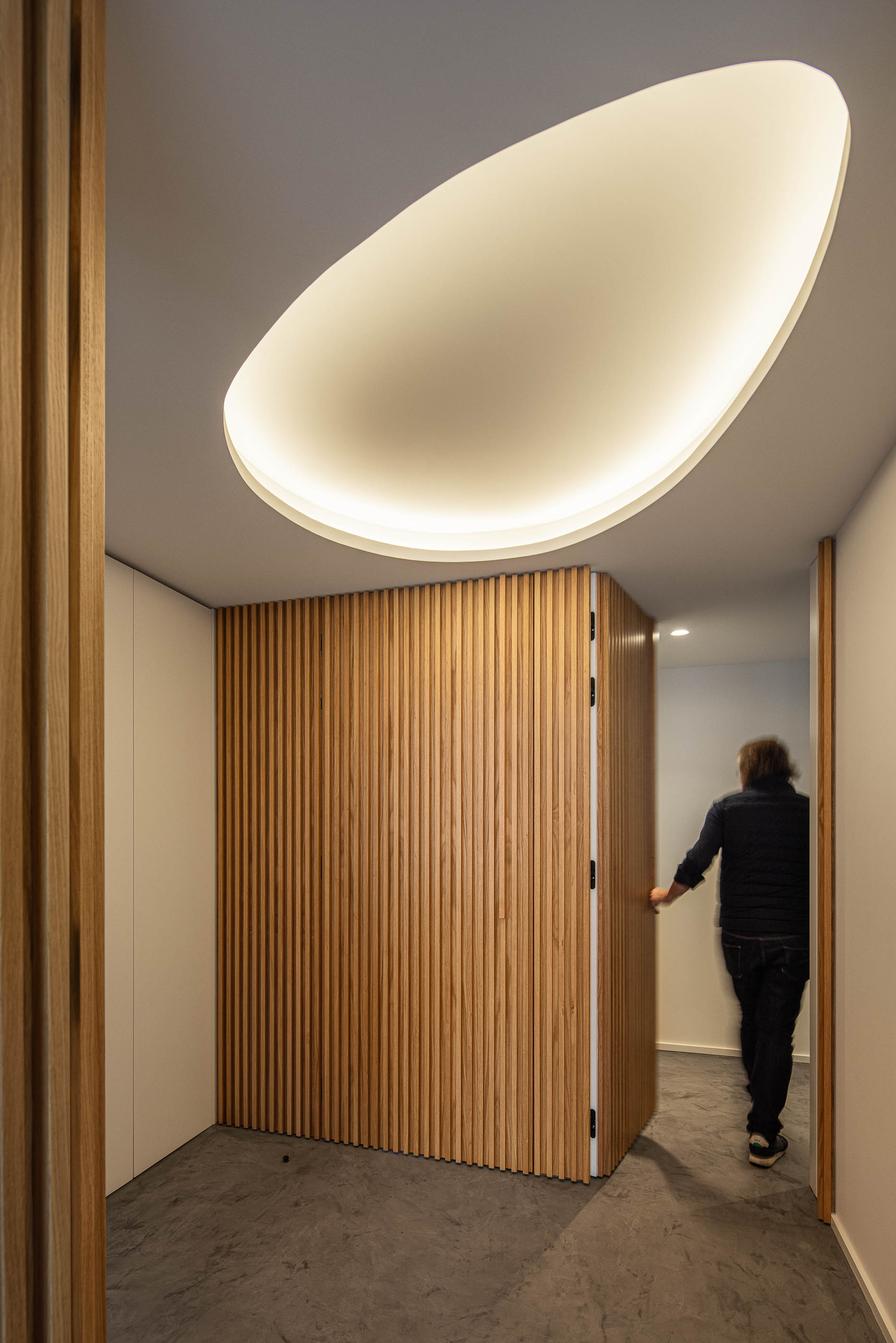 Corredor: Porta pivotante em ripas de madeira clara, armário branco e teto com candeeiro oval. — Wave Apartment arquitetura arQB architecture em Póvoa de Varzim