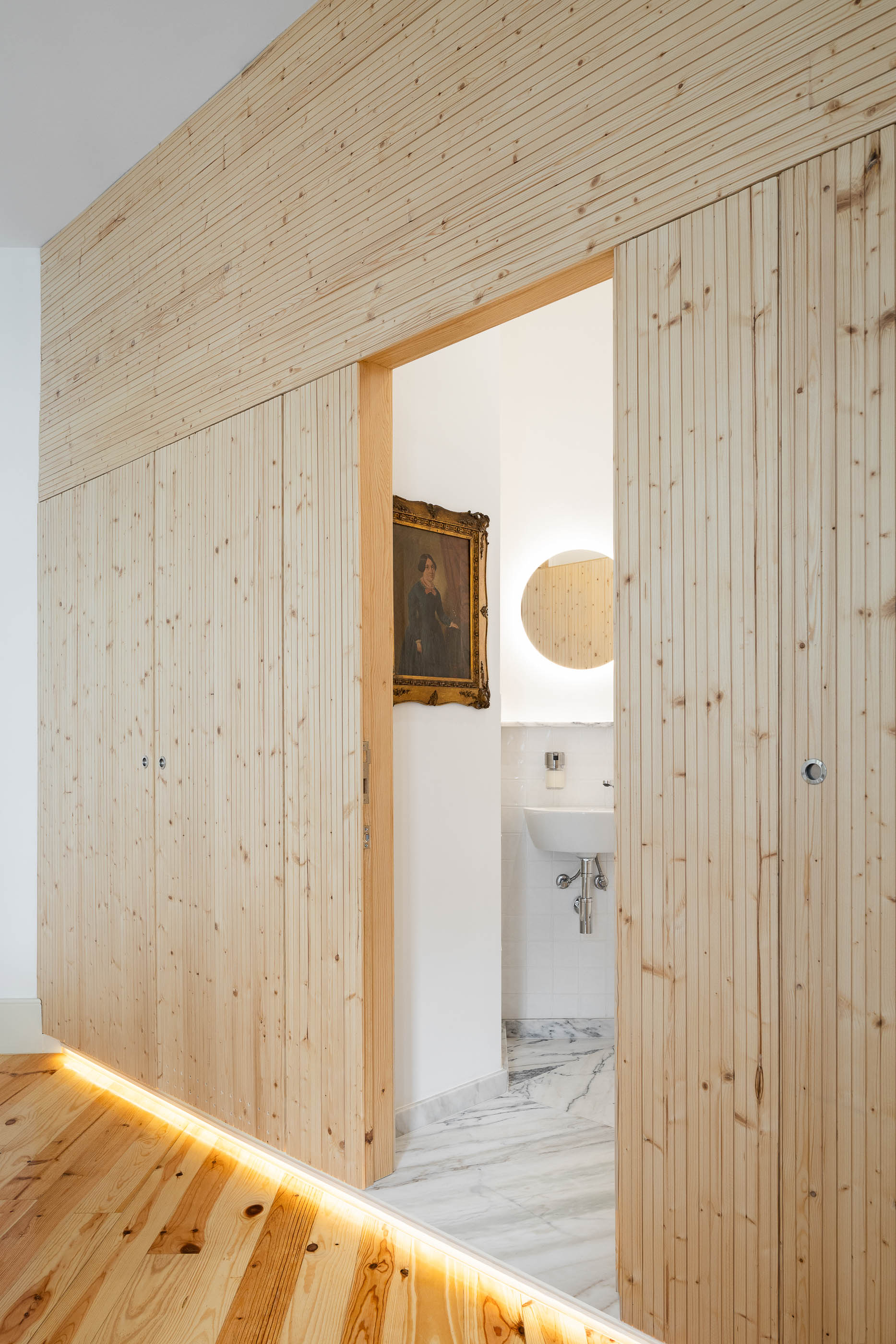 Quarto de dormir com roupeiro em madeira clara e casa de banho adjacente com lavatório branco e espelho circular. — Casa Cedofeita arquitetura Paulo Moreira Architectures em Porto