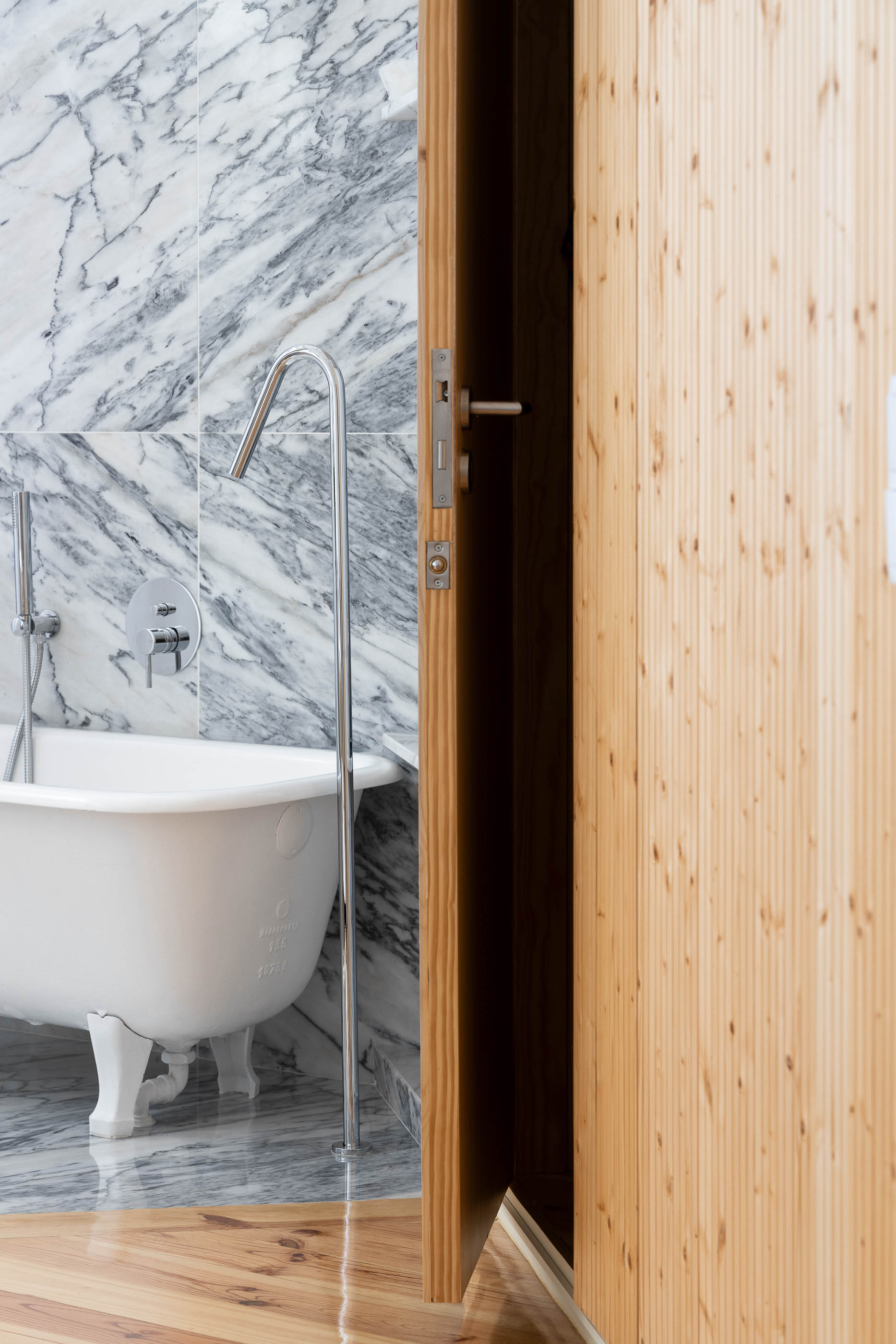 Casa de banho com banheira de pés, revestimento em mármore e porta em madeira clara. — Casa Cedofeita arquitetura Paulo Moreira Architectures em Porto