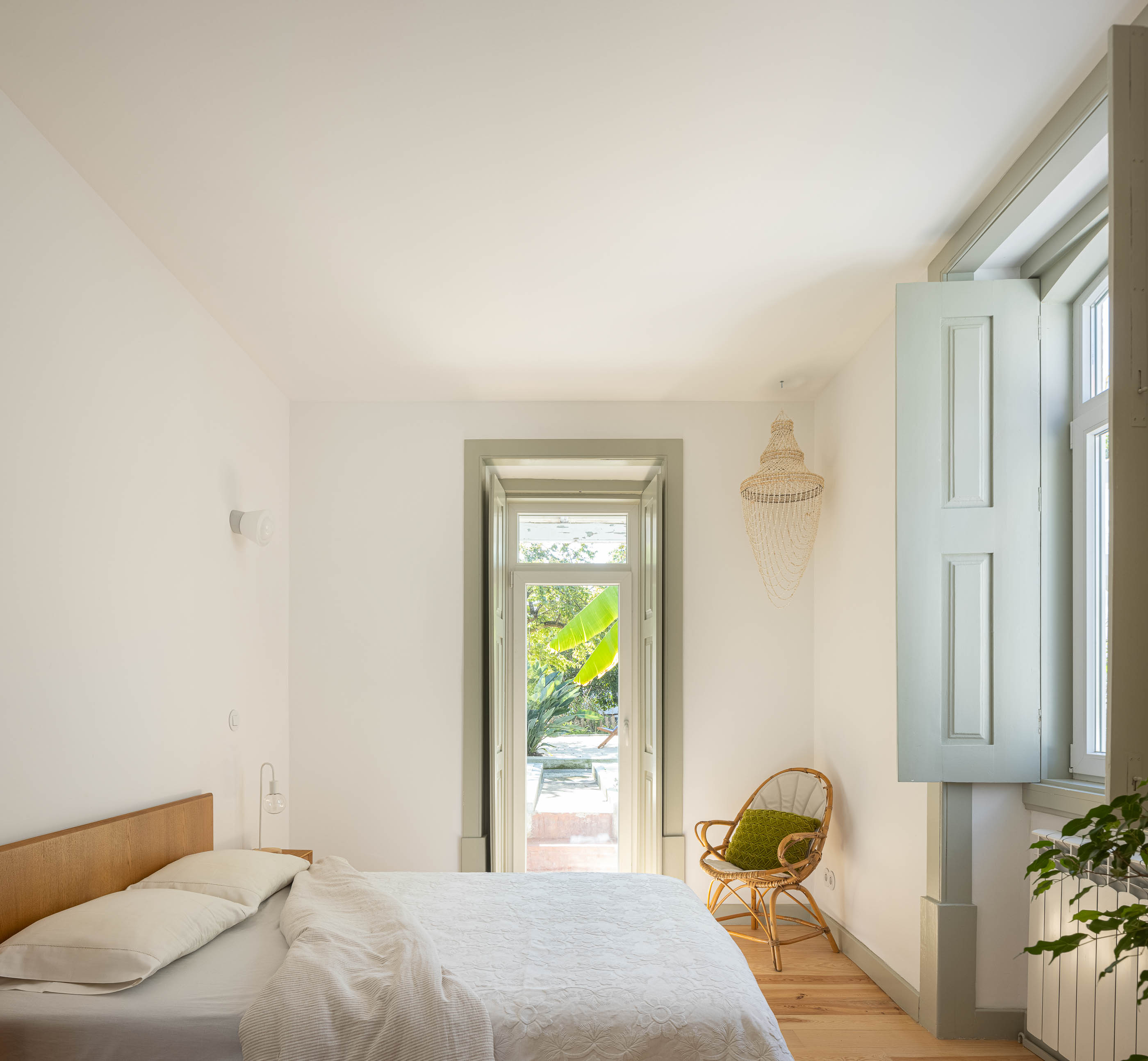 Quarto: Cama de casal com roupa branca, cadeira de vime, candeeiro de teto artesanal e acesso ao exterior. Pavimento em madeira. — Casa em Bencanta arquitetura Filipe Pina Arquitectura em Coimbra