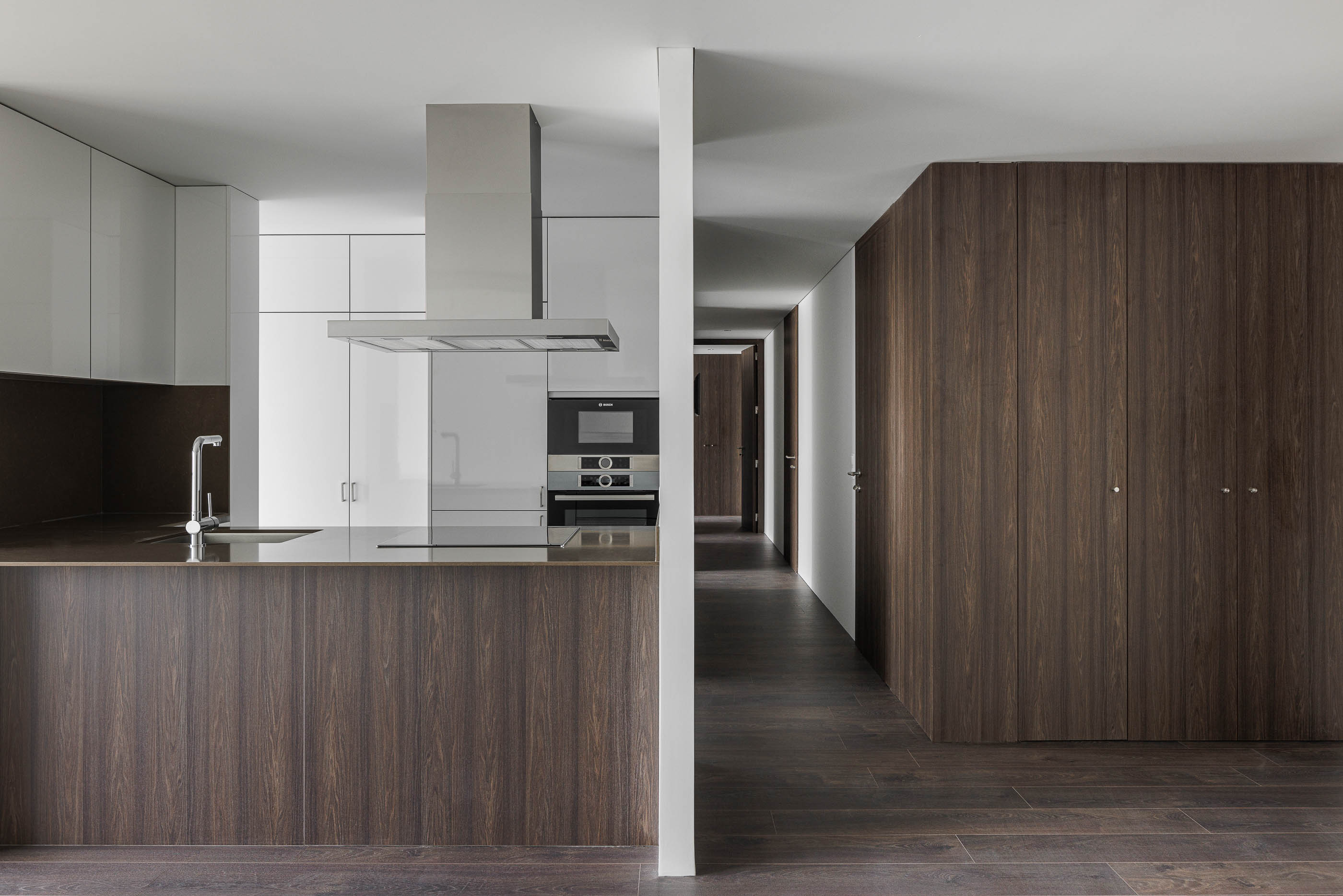 Cozinha: Combinação de armários brancos e revestimento em madeira escura, com ilha central. — Evolution Apartments Housing arquitetura António Paulo Marques, Arquiteto e Associados em Matosinhos