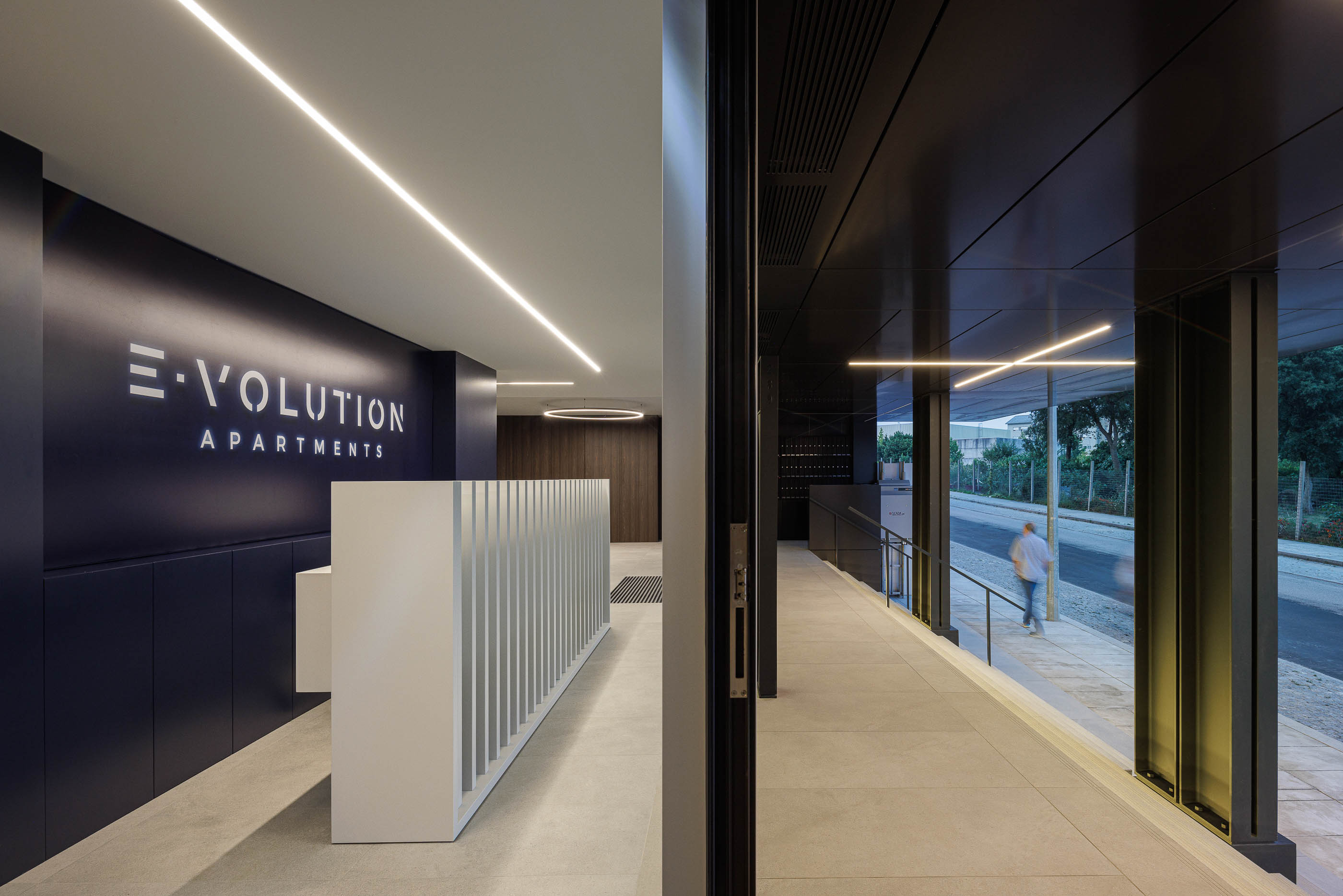 Recepção: Balcão branco com iluminação linear e parede azul escura com o nome do edifício. — Evolution Apartments Housing arquitetura António Paulo Marques, Arquiteto e Associados em Matosinhos