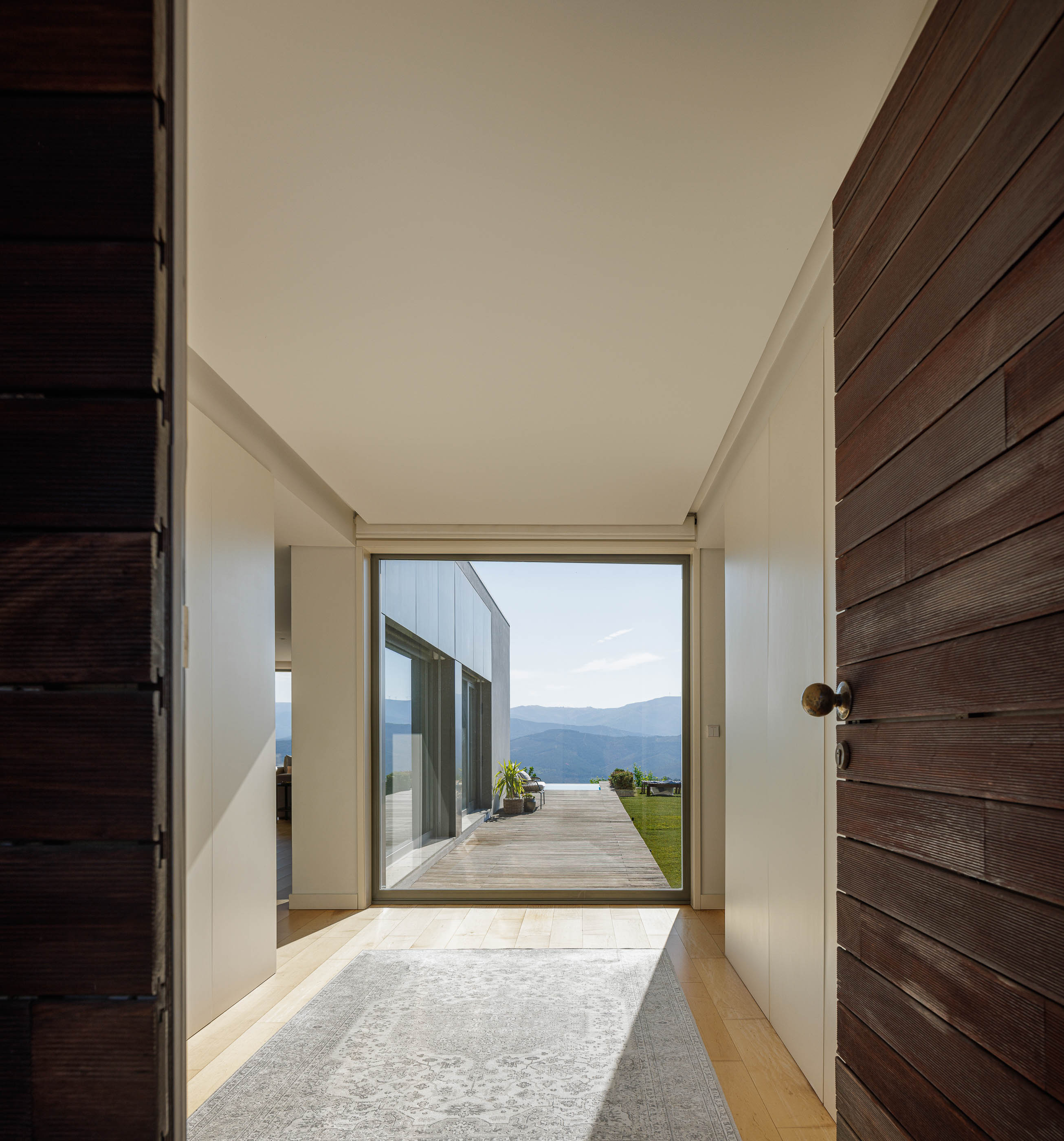Entrada: Porta de madeira escura, pavimento de madeira clara e alcatifa. Vista para o exterior com deck e paisagem montanhosa. — Casa do Alto arquitetura Jorge Martins arquiteto em Celorico de Basto