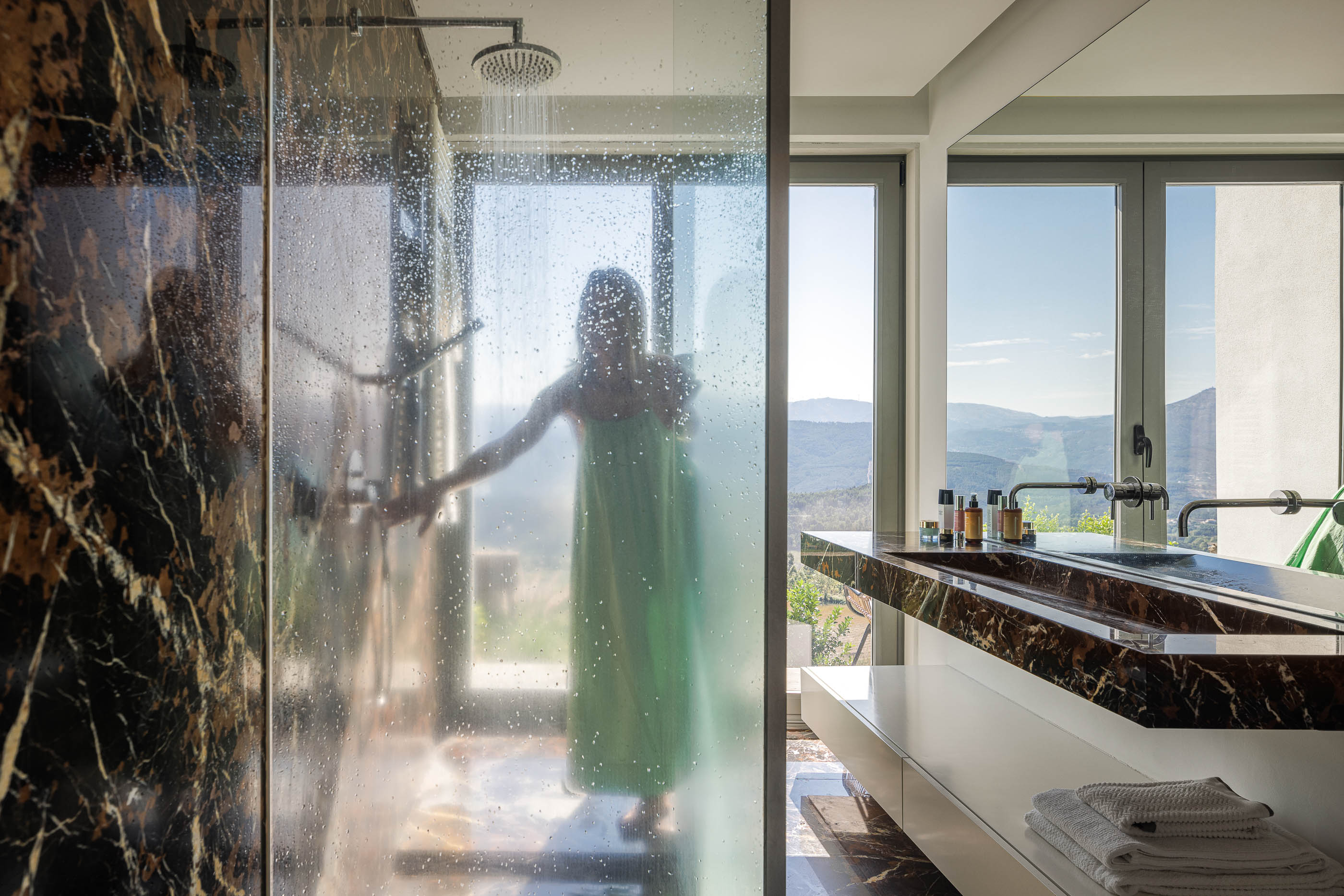 Casa de banho: Cabine de duche em mármore escuro, lavatório embutido no mesmo material, espelho amplo e janela com vista. — Casa do Alto arquitetura Jorge Martins arquiteto em Celorico de Basto