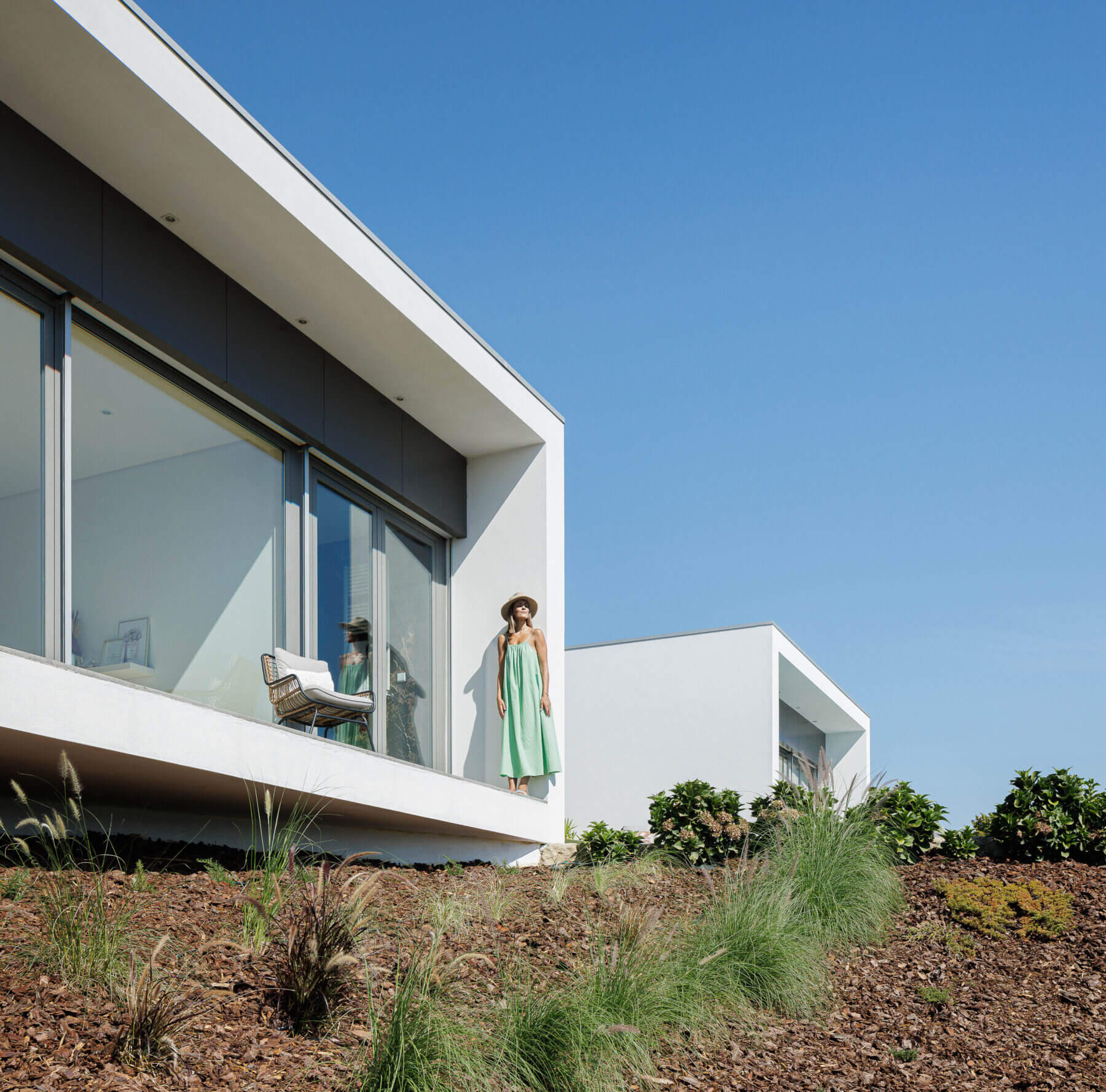 Exterior: Fachada branca com detalhes em cinza, grande janela e varanda com cadeira. Paisagem natural com vegetação rasteira. — Casa do Alto arquitetura Jorge Martins arquiteto em Celorico de Basto