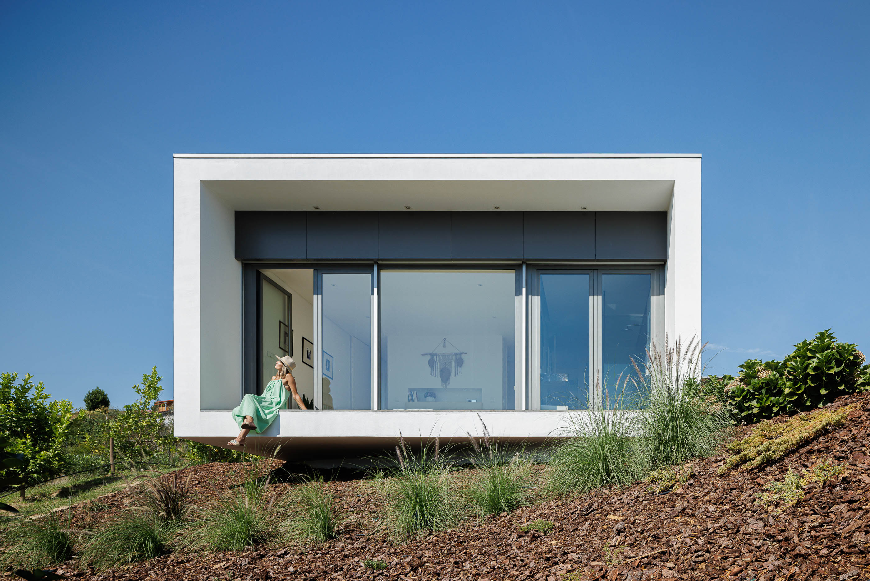 Exterior: Fachada branca com detalhes em cinza, grande janela e varanda. Paisagem natural com vegetação rasteira e vinha. — Casa do Alto arquitetura Jorge Martins arquiteto em Celorico de Basto