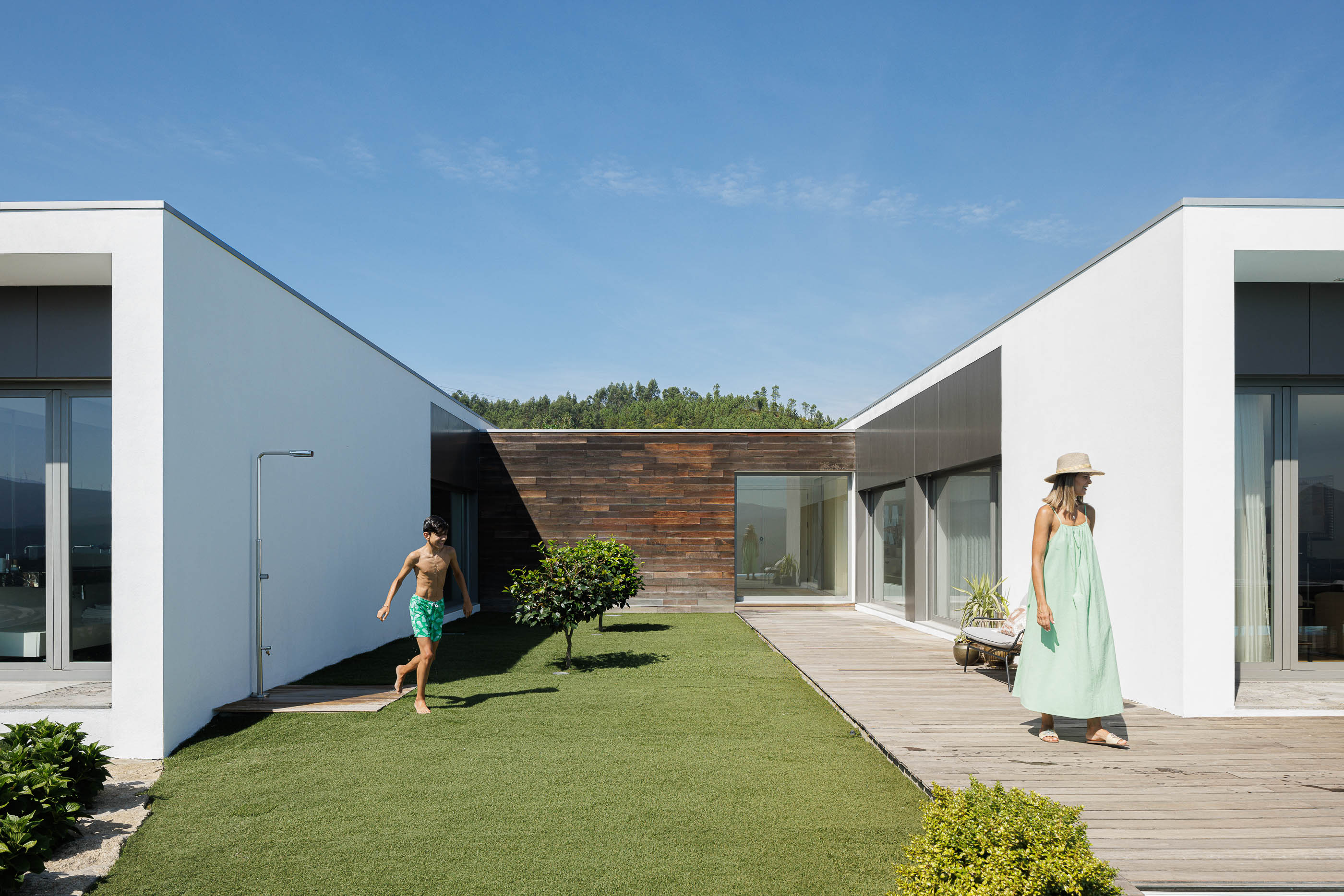 Exterior: Vista do pátio interior com relvado, deck de madeira, fachada em madeira e volumes brancos da casa. — Casa do Alto arquitetura Jorge Martins arquiteto em Celorico de Basto