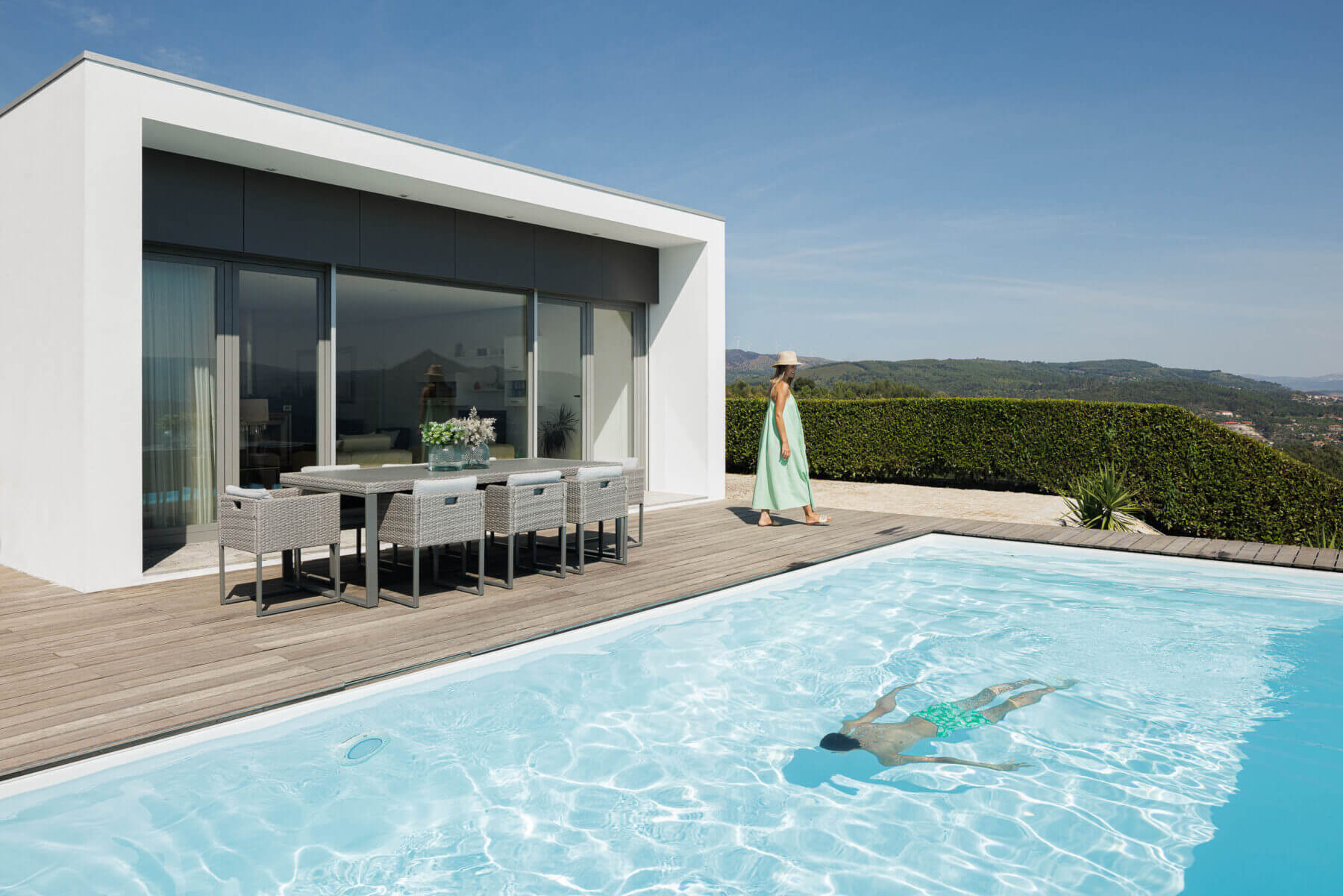 Exterior: Piscina com deck de madeira, mesa de refeições e vista panorâmica. Fachada branca da casa com grandes janelas. — Casa do Alto arquitetura Jorge Martins arquiteto em Celorico de Basto