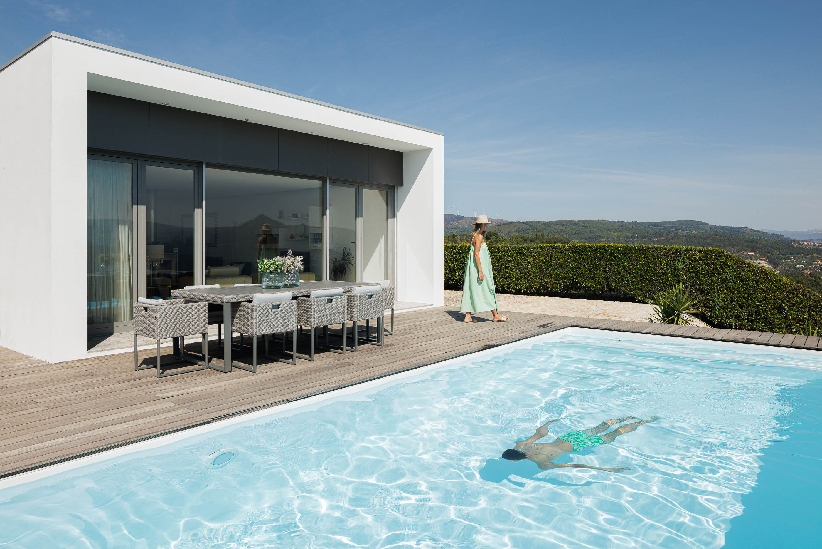 Exterior: Piscina com deck de madeira, mesa de refeições e vista panorâmica. Fachada branca da casa com grandes janelas. — Casa do Alto arquitetura Jorge Martins arquiteto em Celorico de Basto