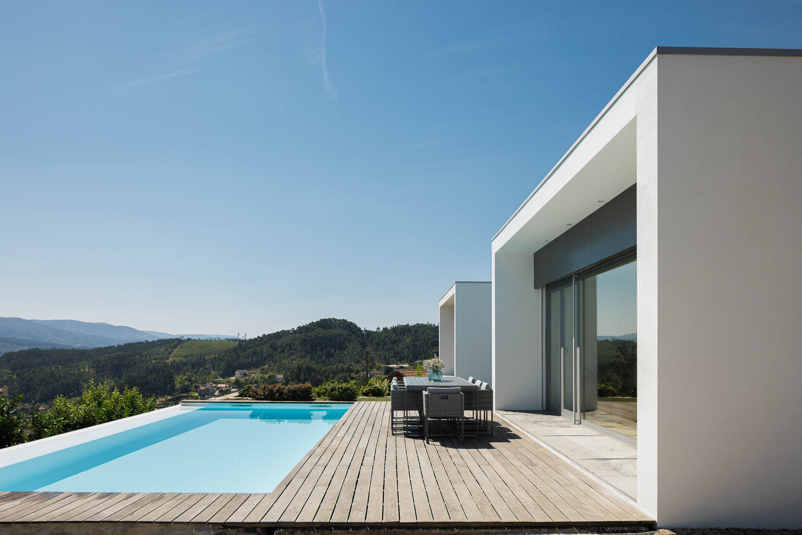 Exterior: Piscina com deck de madeira, mesa de refeições e vista panorâmica. Fachada branca da casa com grandes janelas. — Casa do Alto arquitetura Jorge Martins arquiteto em Celorico de Basto