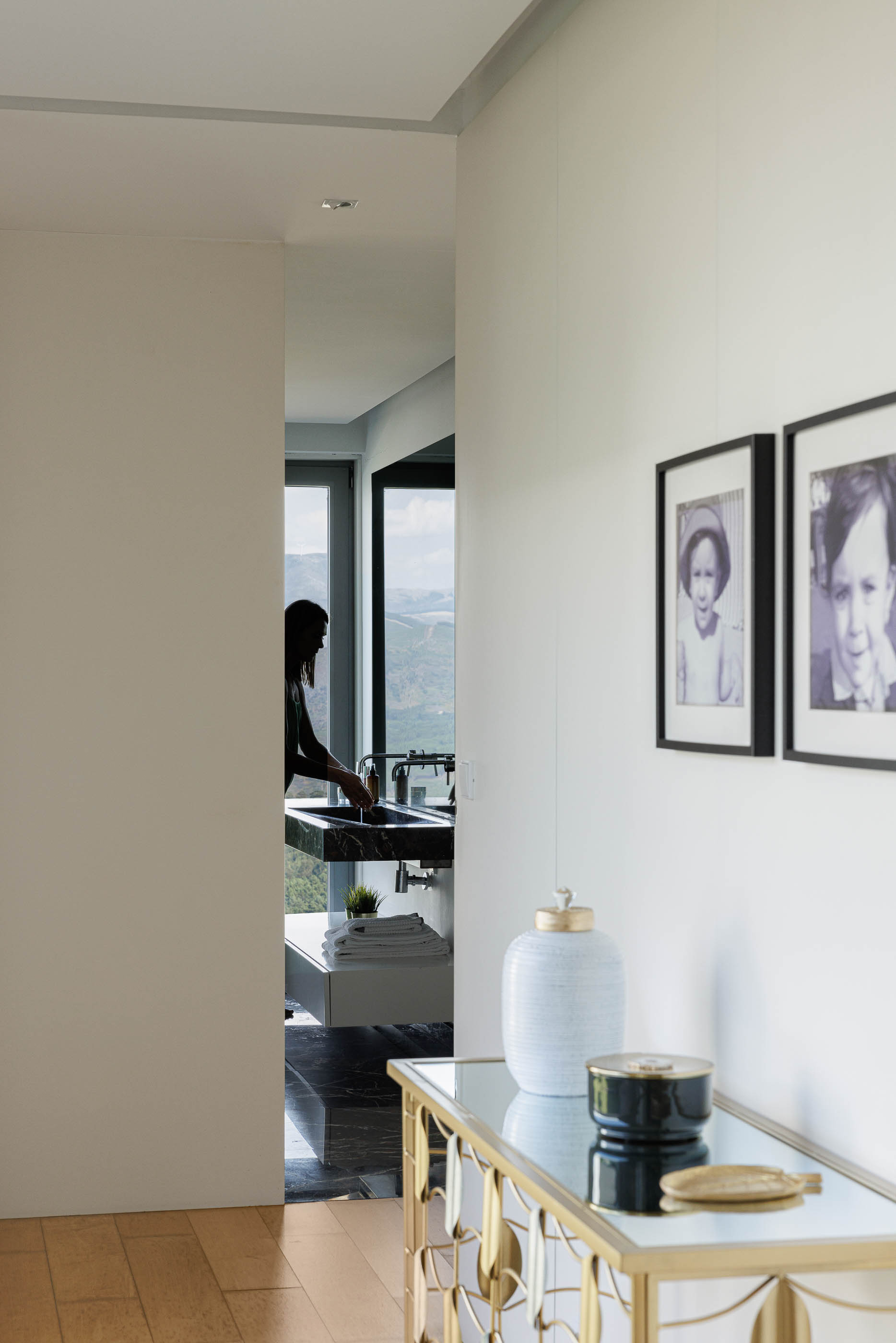 Corredor: Consola dourada com objetos decorativos, fotografias emolduradas e vista para a casa de banho com lavatório em mármore. — Casa do Alto arquitetura Jorge Martins arquiteto em Celorico de Basto