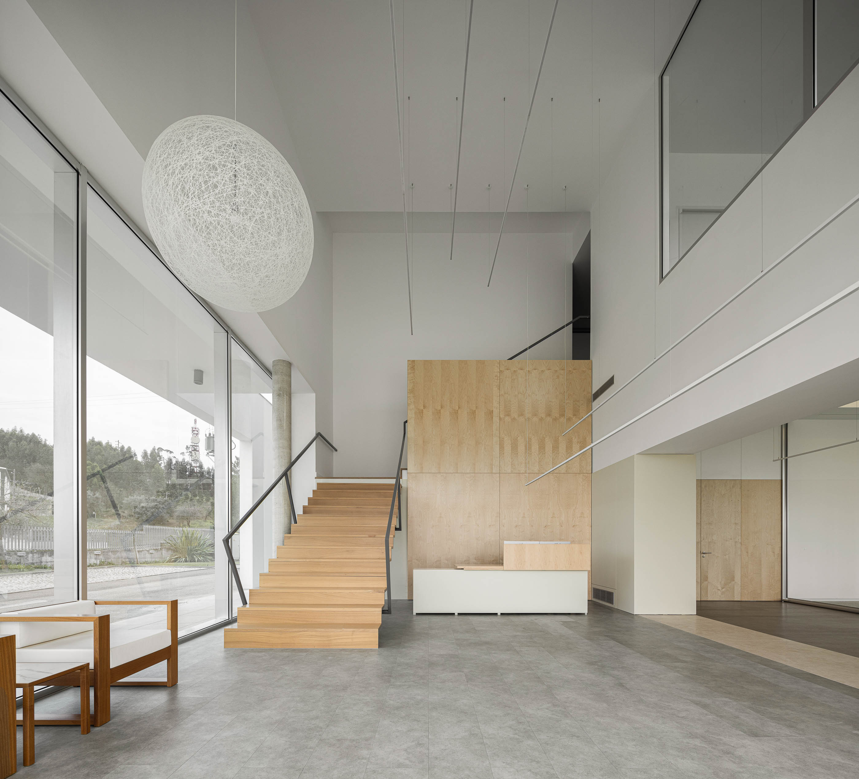 Hall de entrada com escadas em madeira, candeeiro esférico e pavimento cinzento. Estilo minimalista com grandes janelas. — Edifício Sede da PRF arquitetura Impare Arquitectura em Leiria