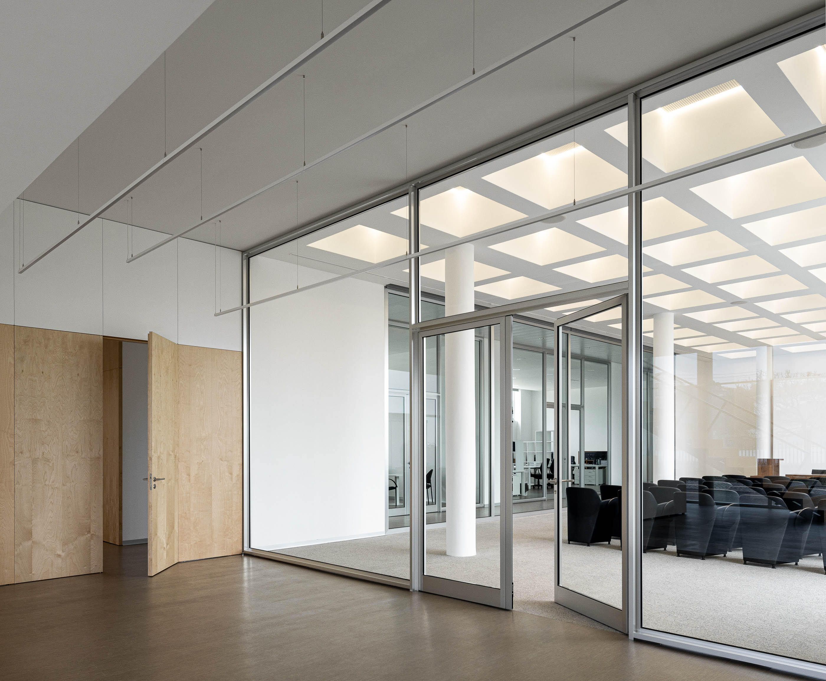 Sala de reuniões com cadeiras pretas, paredes de vidro e pavimento castanho. Iluminação em grelha no teto. — Edifício Sede da PRF arquitetura Impare Arquitectura em Leiria