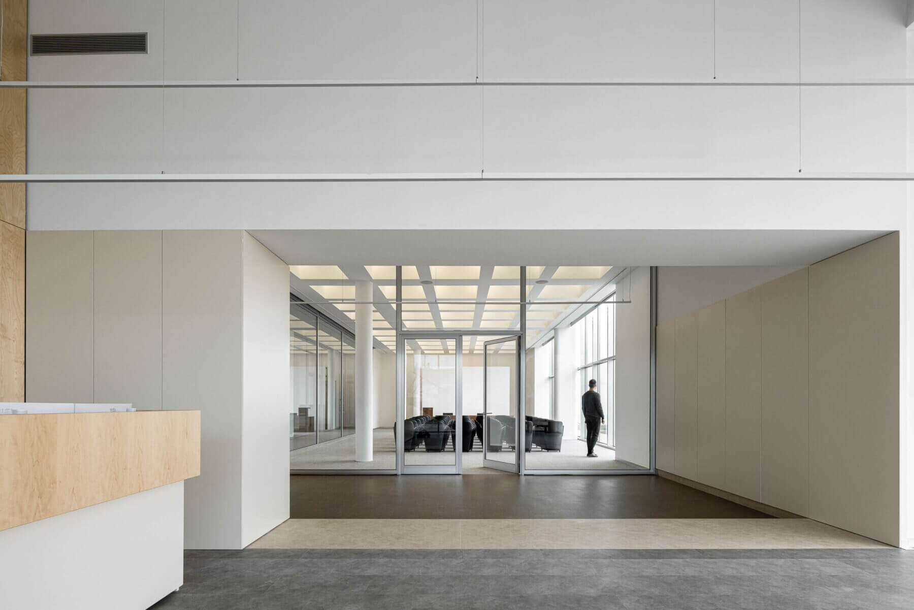 Hall de entrada com balcão em madeira clara e acesso à sala de reuniões envidraçada. Pavimento em tons neutros. — Edifício Sede da PRF arquitetura Impare Arquitectura em Leiria