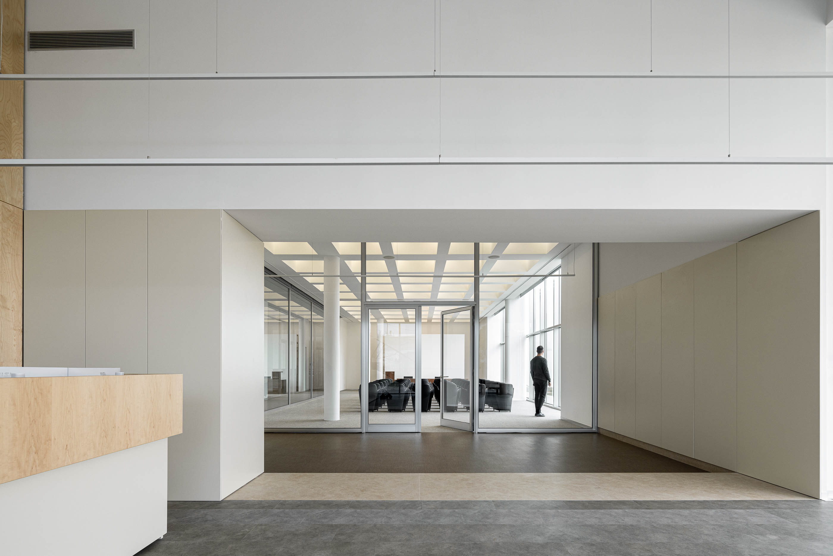 Hall de entrada com balcão em madeira clara e acesso à sala de reuniões envidraçada. Pavimento em tons neutros. — Edifício Sede da PRF arquitetura Impare Arquitectura em Leiria
