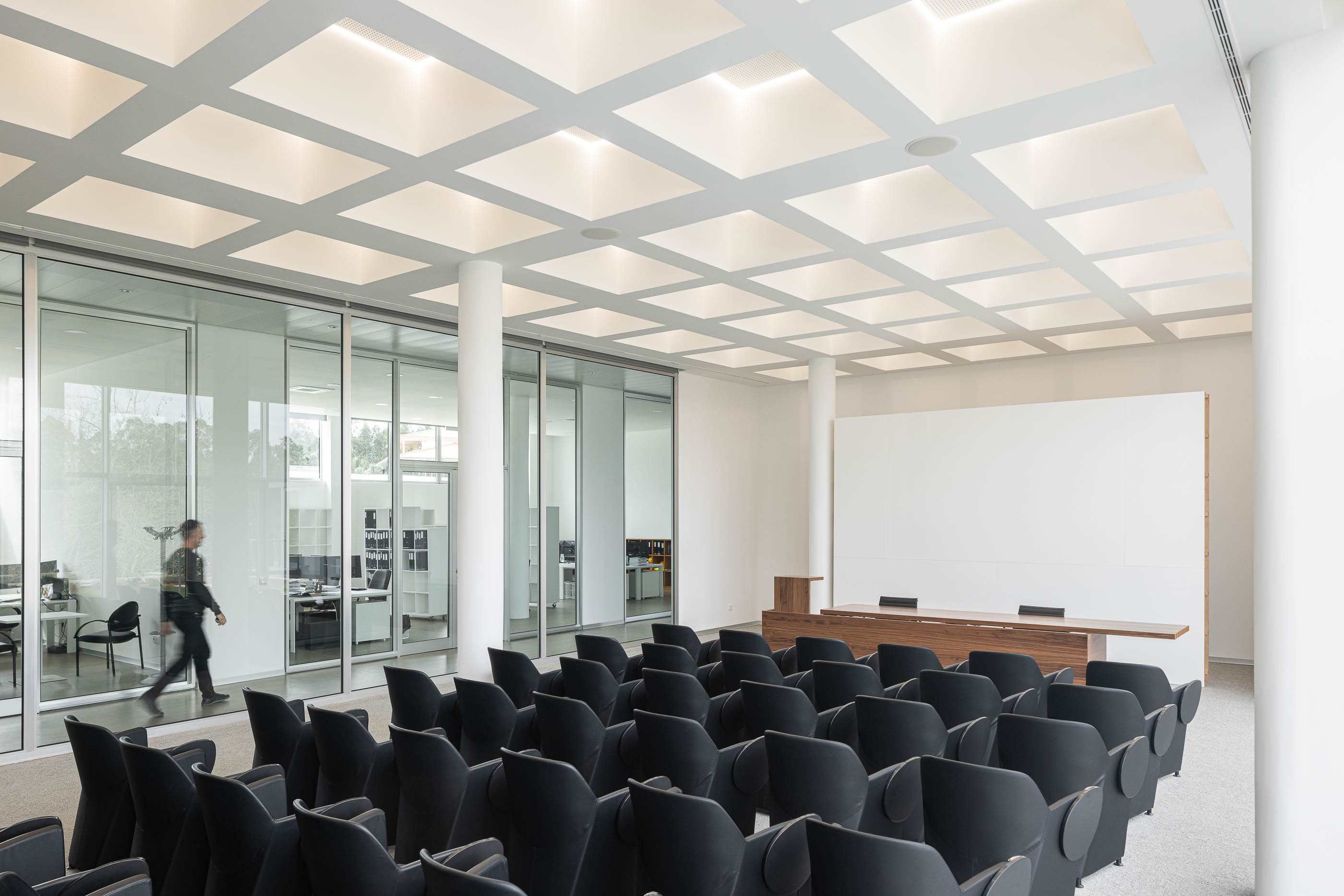 Sala de conferências com cadeiras pretas, mesa de oradores em madeira e paredes de vidro. Pavimento alcatifado cinzento. — Edifício Sede da PRF arquitetura Impare Arquitectura em Leiria