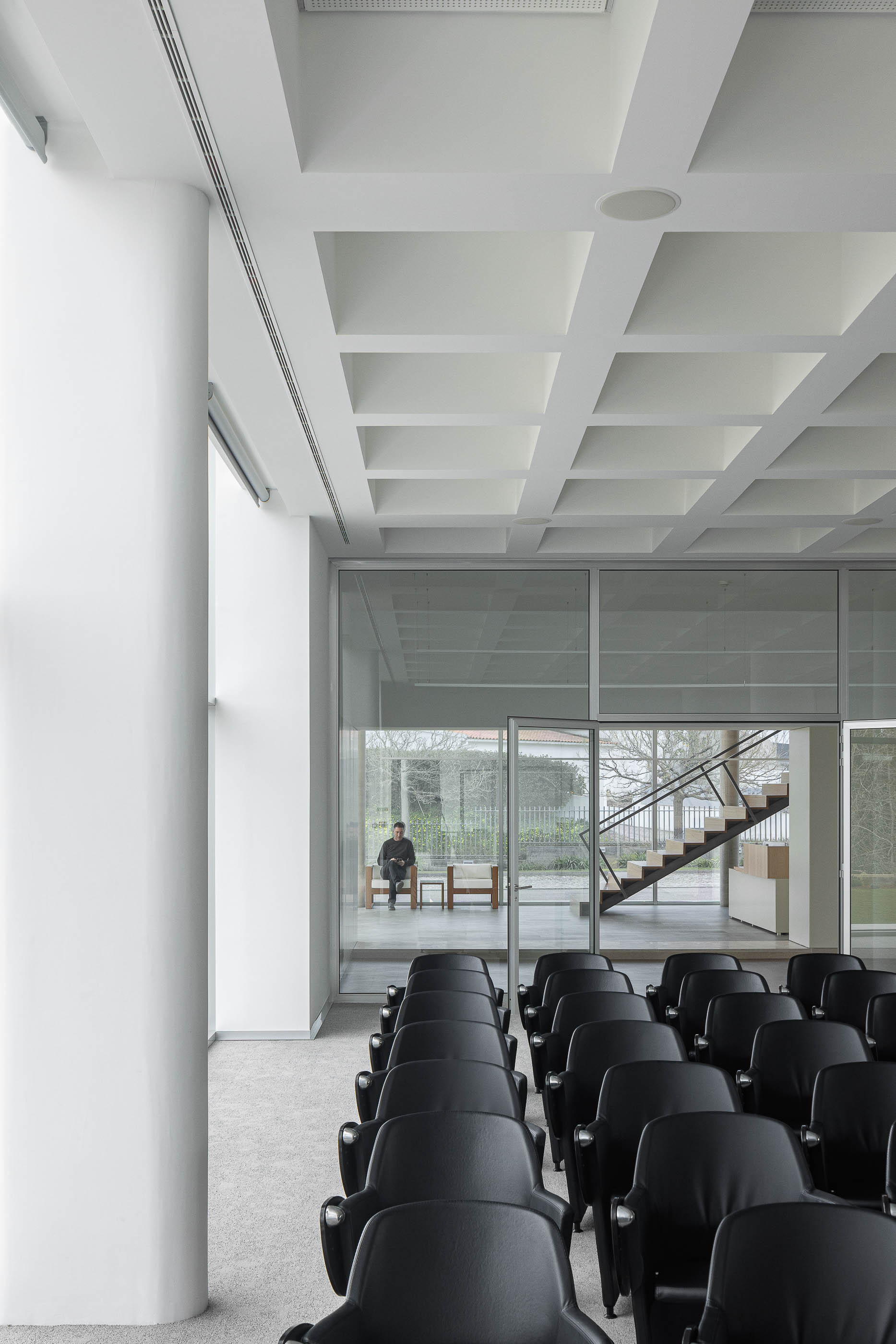 Sala de conferências com cadeiras pretas, colunas brancas e vista para o exterior. Pavimento alcatifado cinzento. — Edifício Sede da PRF arquitetura Impare Arquitectura em Leiria