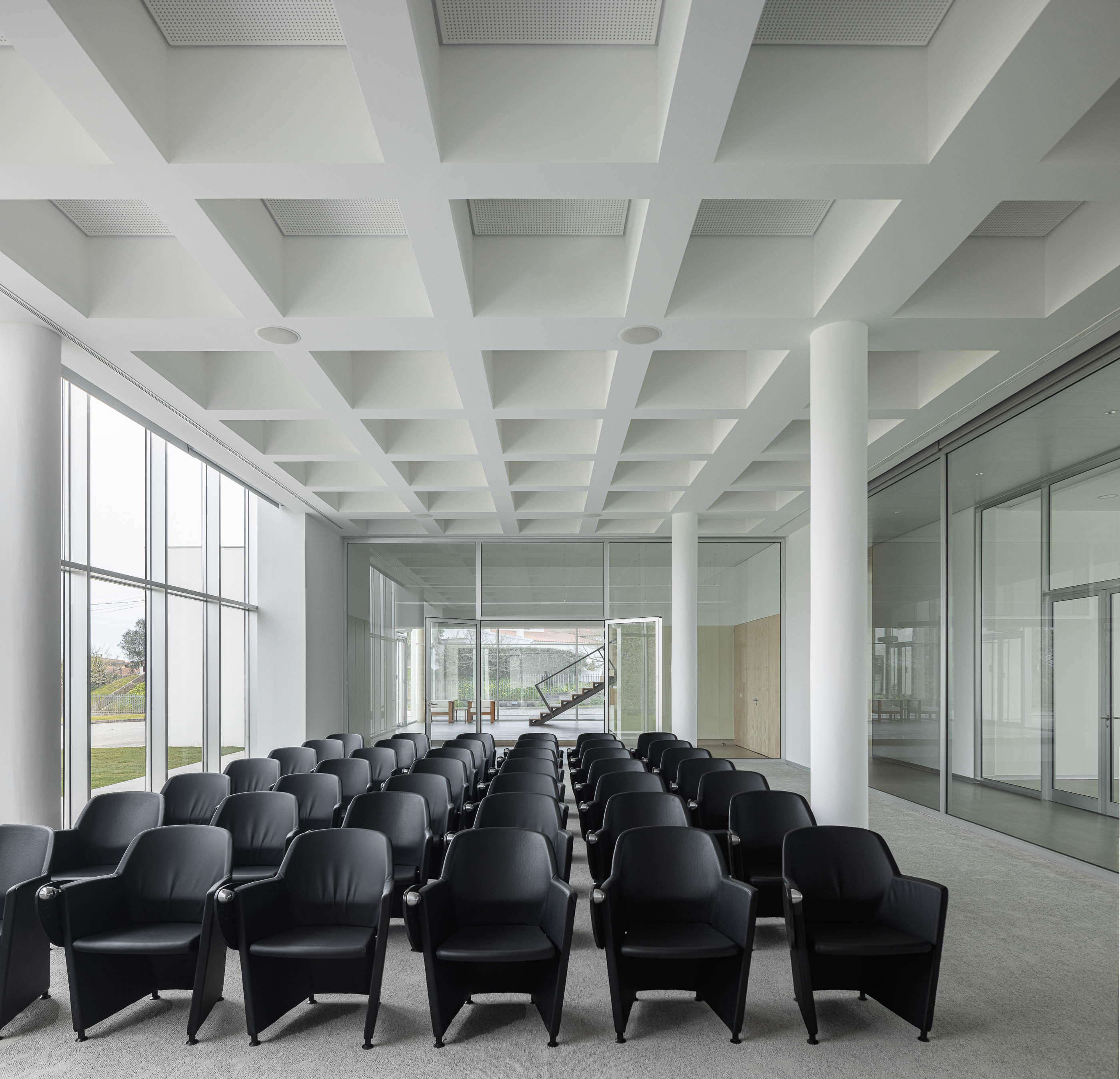 Sala de conferências com cadeiras pretas, paredes de vidro e iluminação embutida no teto. Pavimento alcatifado cinzento. — Edifício Sede da PRF arquitetura Impare Arquitectura em Leiria