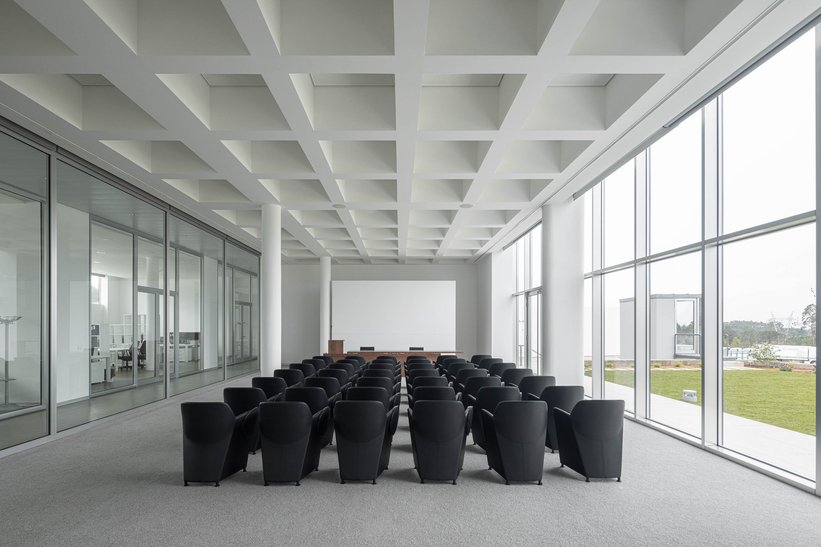 Sala de conferências com cadeiras pretas, paredes de vidro e vista para o exterior. Pavimento alcatifado cinzento. — Edifício Sede da PRF arquitetura Impare Arquitectura em Leiria