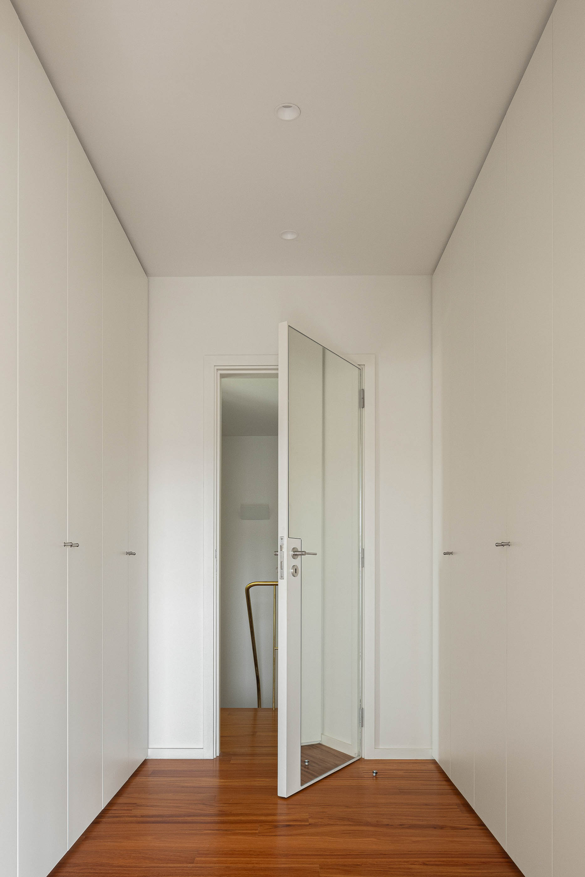 Closet: Corredor com roupeiros brancos embutidos e pavimento em madeira. Porta aberta para as escadas. — Casa IMLA arquitetura Luppa em Porto