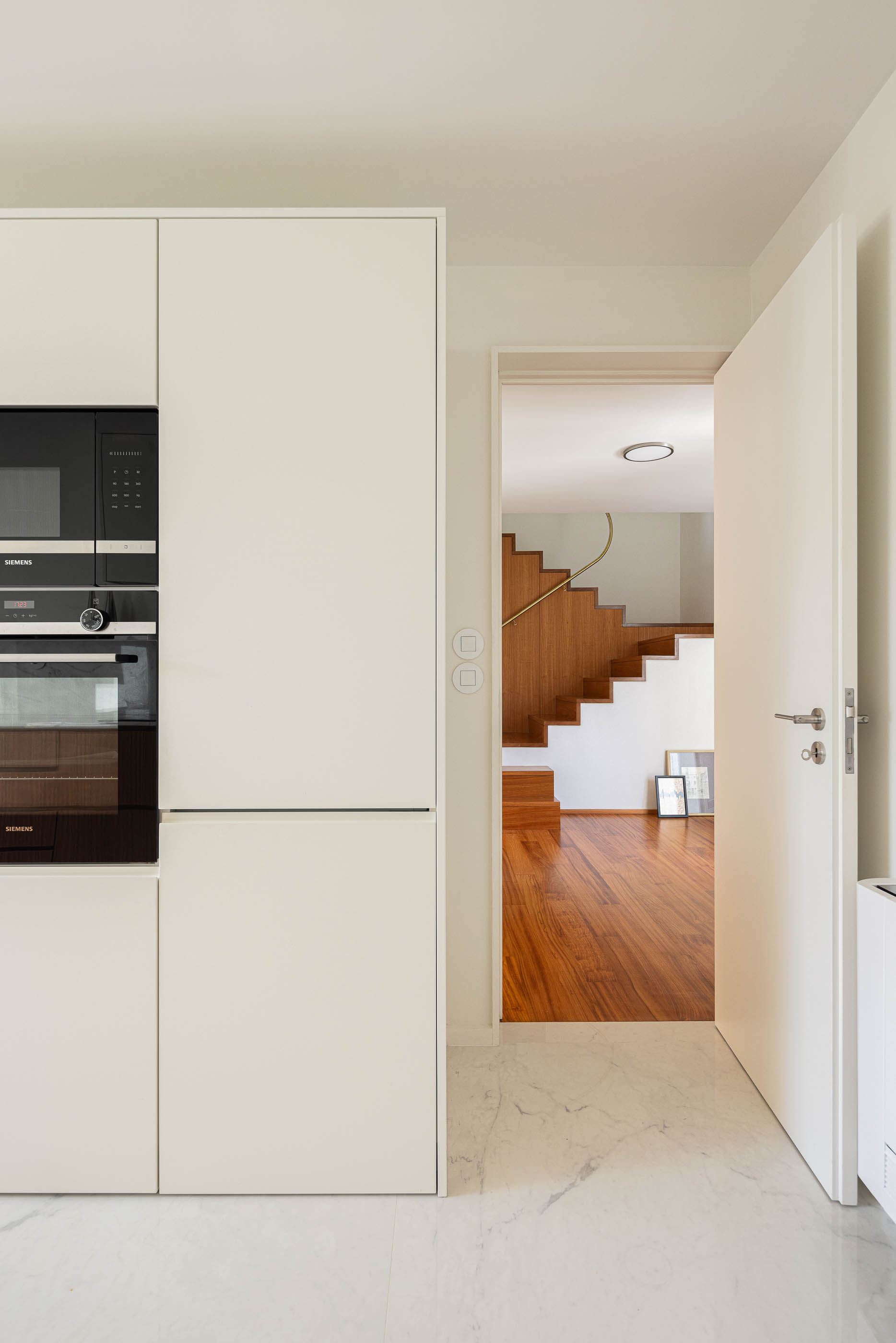 Cozinha: Armários brancos embutidos com eletrodomésticos pretos, porta aberta para as escadas. — Casa IMLA arquitetura Luppa em Porto
