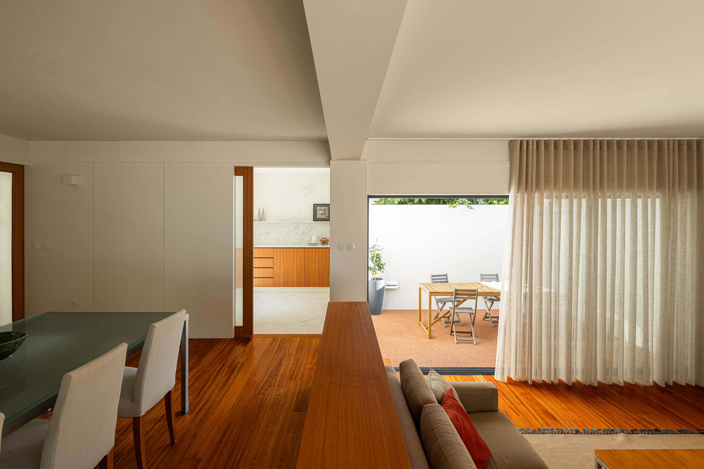 Sala de estar: Vista da sala de jantar, cozinha e pátio, com pavimento em madeira e cortinas claras. — Casa IMLA arquitetura Luppa em Porto