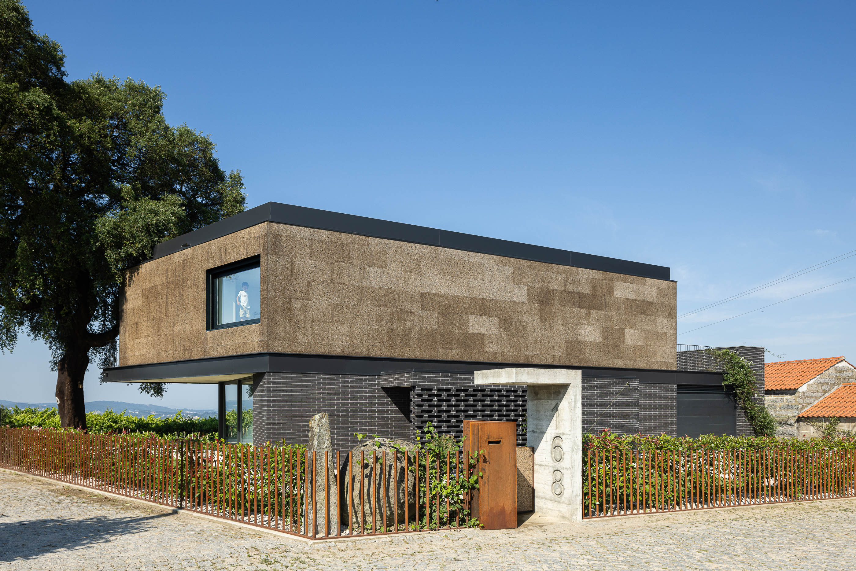 Fachada: Vista exterior da casa com revestimento de cortiça e tijolo preto, jardim com vedação metálica e sobreiro. — Casa do Sobreiro arquitetura Skemba - Arquitectura e Engenharia em Amarante