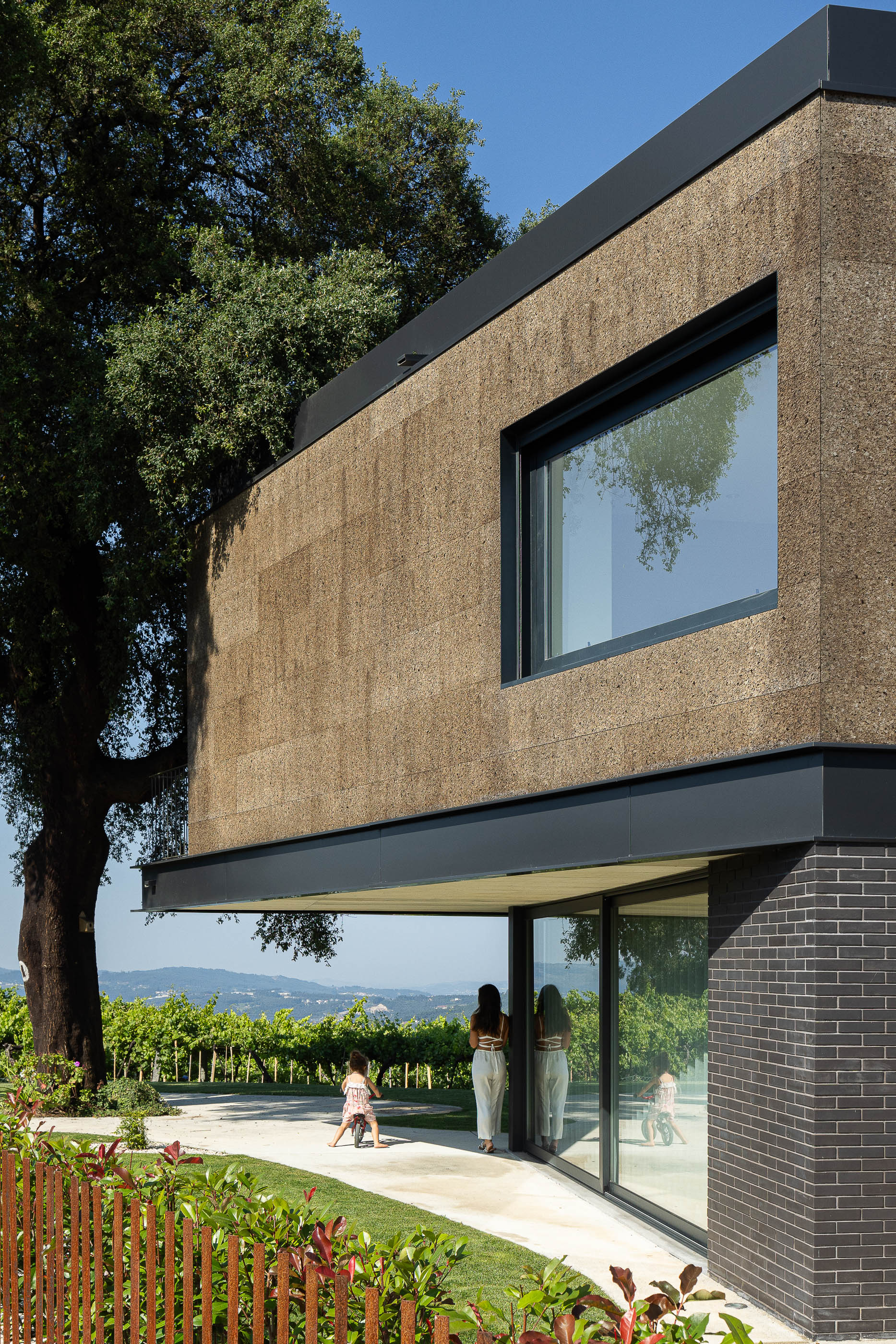 Jardim: Vista da fachada com revestimento de cortiça e tijolo, com janelas amplas e vinhas ao fundo. — Casa do Sobreiro arquitetura Skemba - Arquitectura e Engenharia em Amarante
