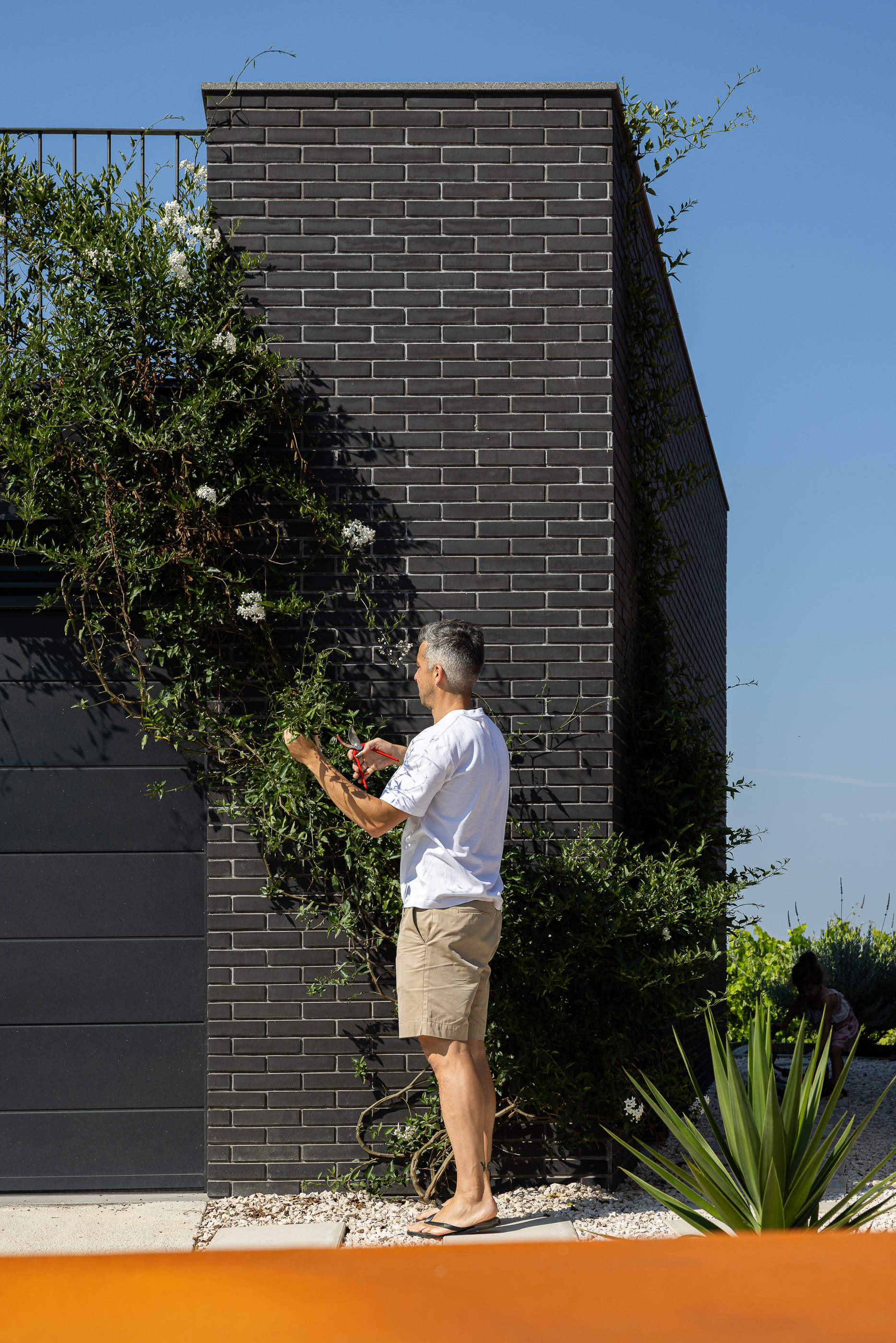 Exterior: Parede de tijolo preto com vegetação trepadeira e homem a podar as plantas. — Casa do Sobreiro arquitetura Skemba - Arquitectura e Engenharia em Amarante
